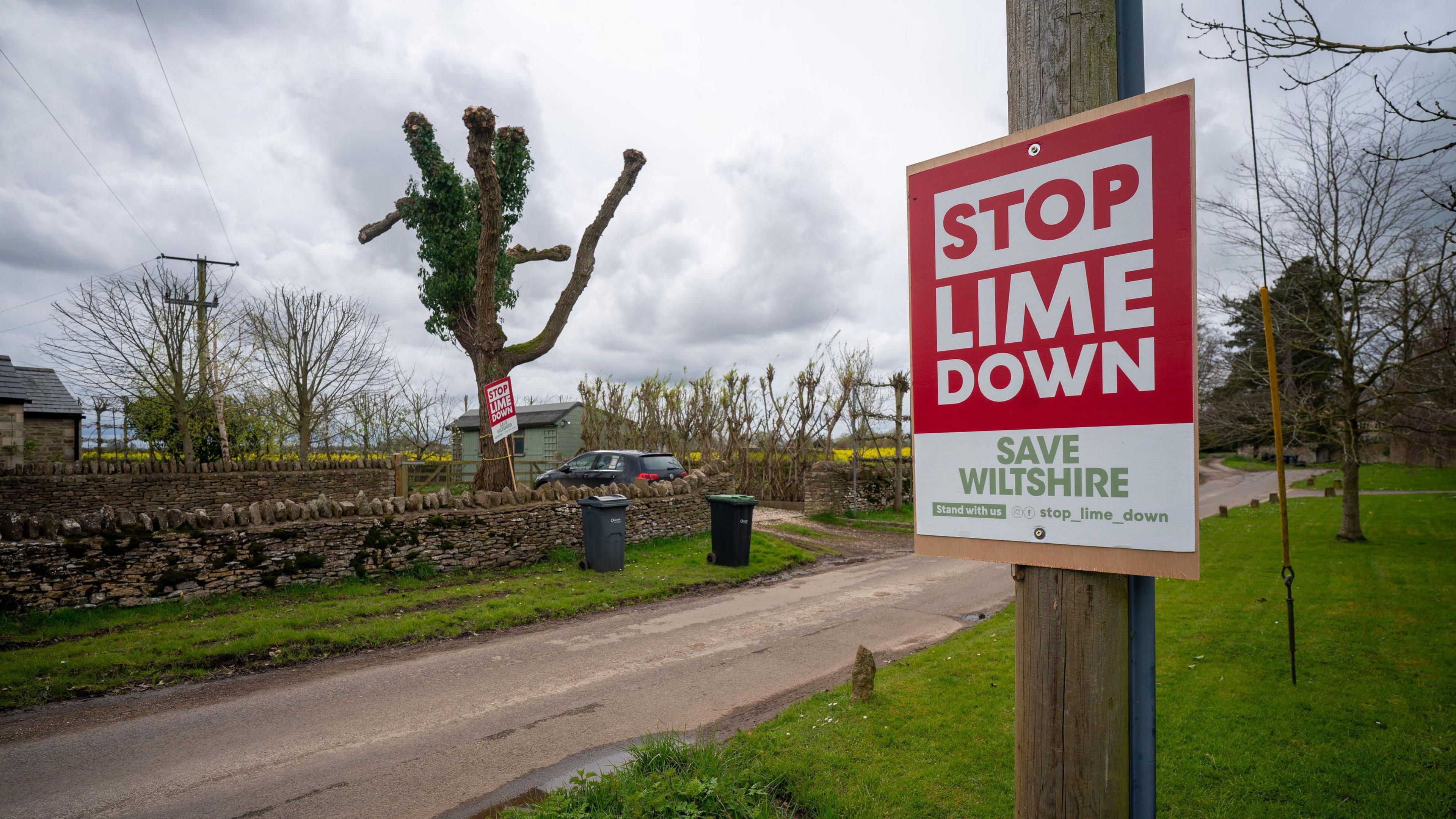 A sign pinned to a wooden pole reads 'Stop Lime Down Save Wiltshire'  on a country road.