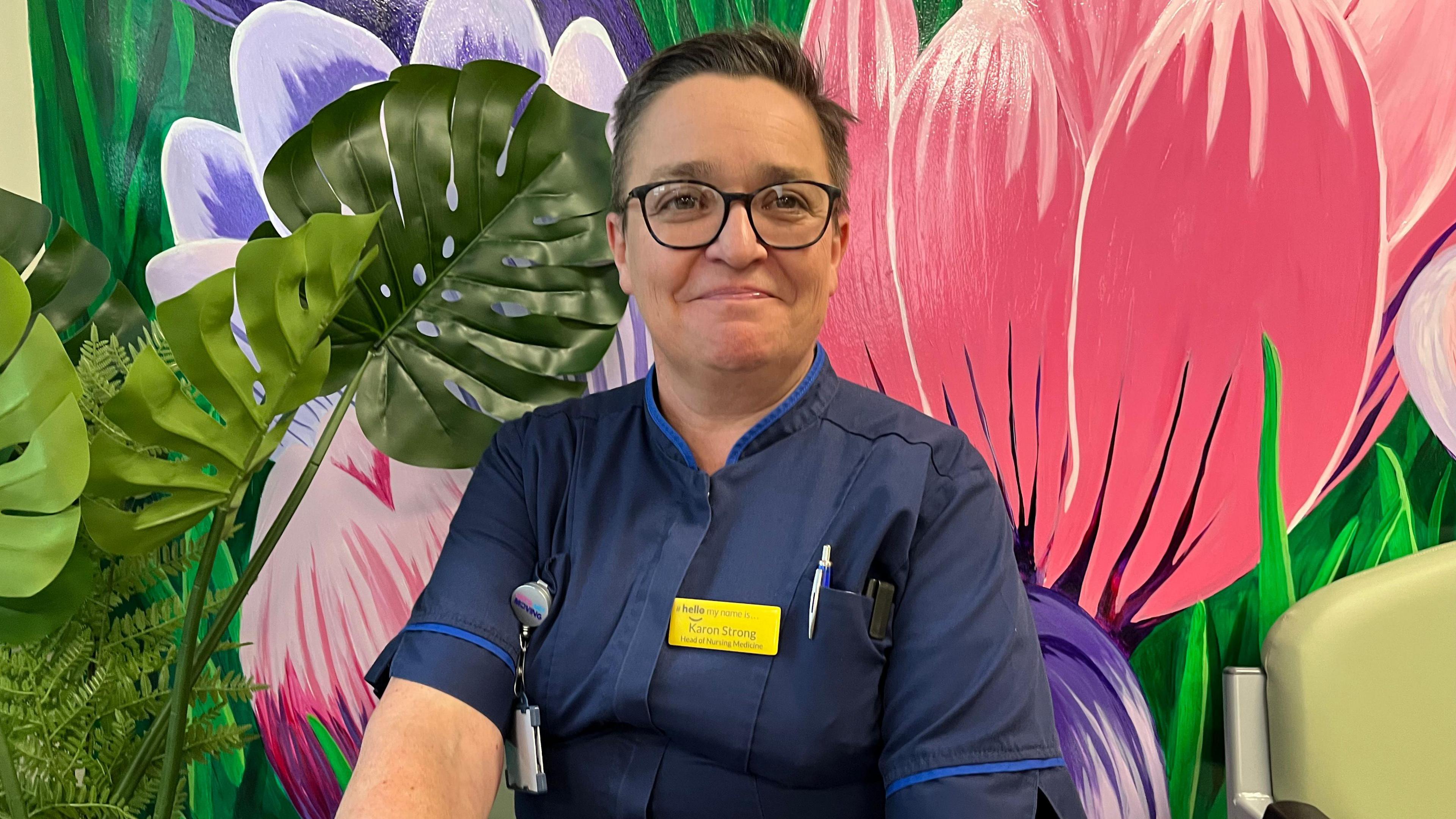 Karon Strong sits in front of a large floral mural. She is wearing blue nurses' scrubs and has short dark hair and wears glasses.