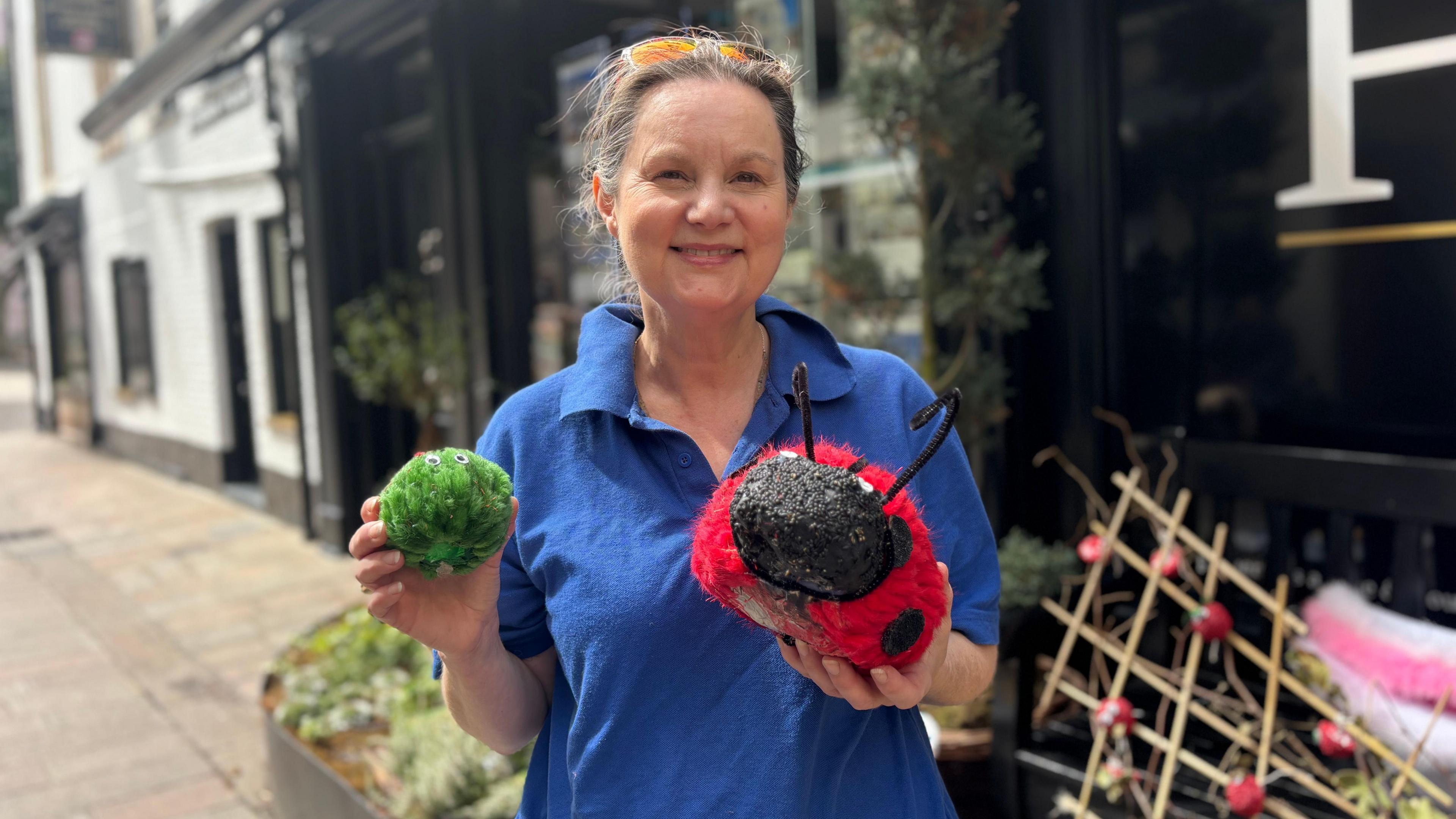 A woman, wearing a blue t-shirt and sunglasses on her head. She is holding a knitted Brussels sprout and knitted ladybird. 