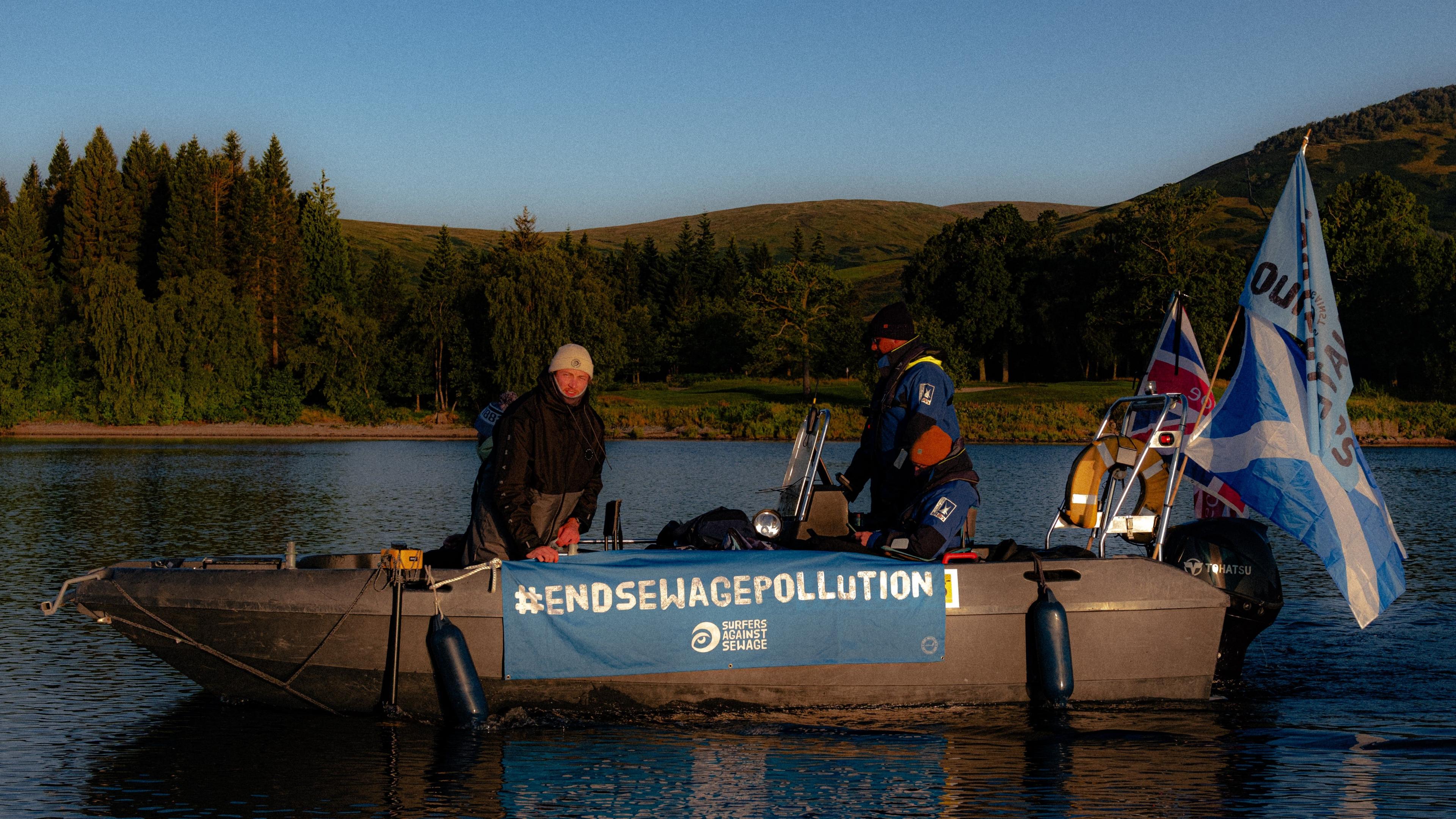 Two people on a boat with banner that says #endsewagepollution with surfers against sewage logo and a lake and landscape in background