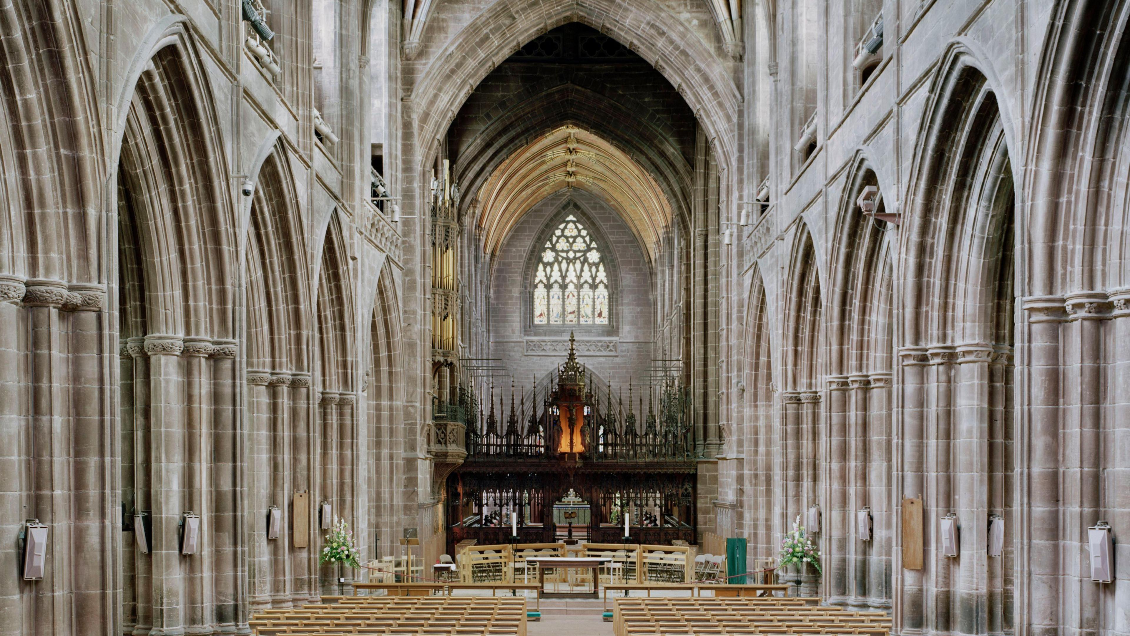 The nave of Chester Cathedral shows light brown benches. There are high walls that are arched and flowers on the side of the stage