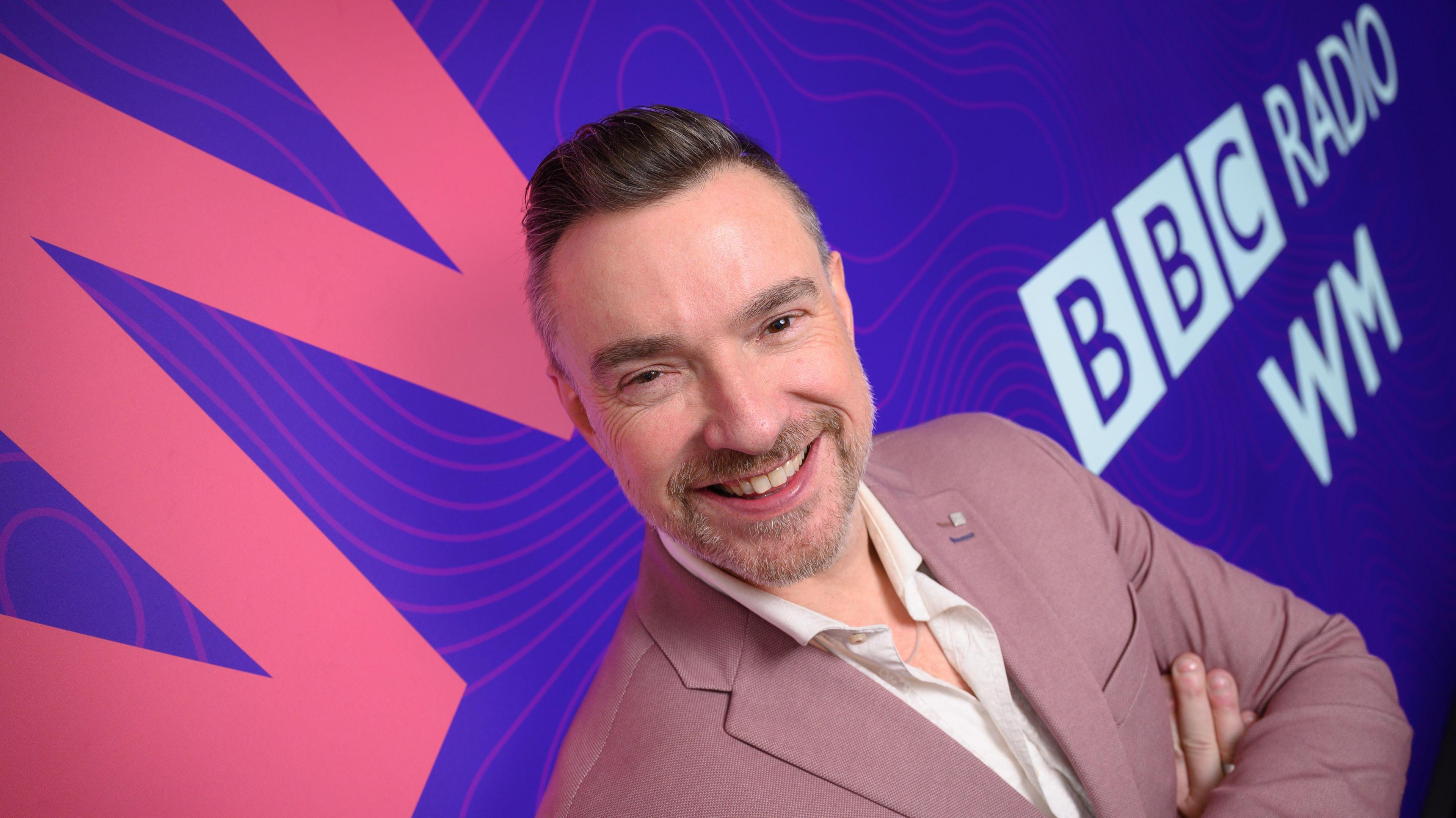 Ed James has his arms crossed and is standing in front of a purple and pink banner that says BBC Radio WM. He has short brown hair and grey facial hair and is wearing a dusty pink suit jacket with white shirt.