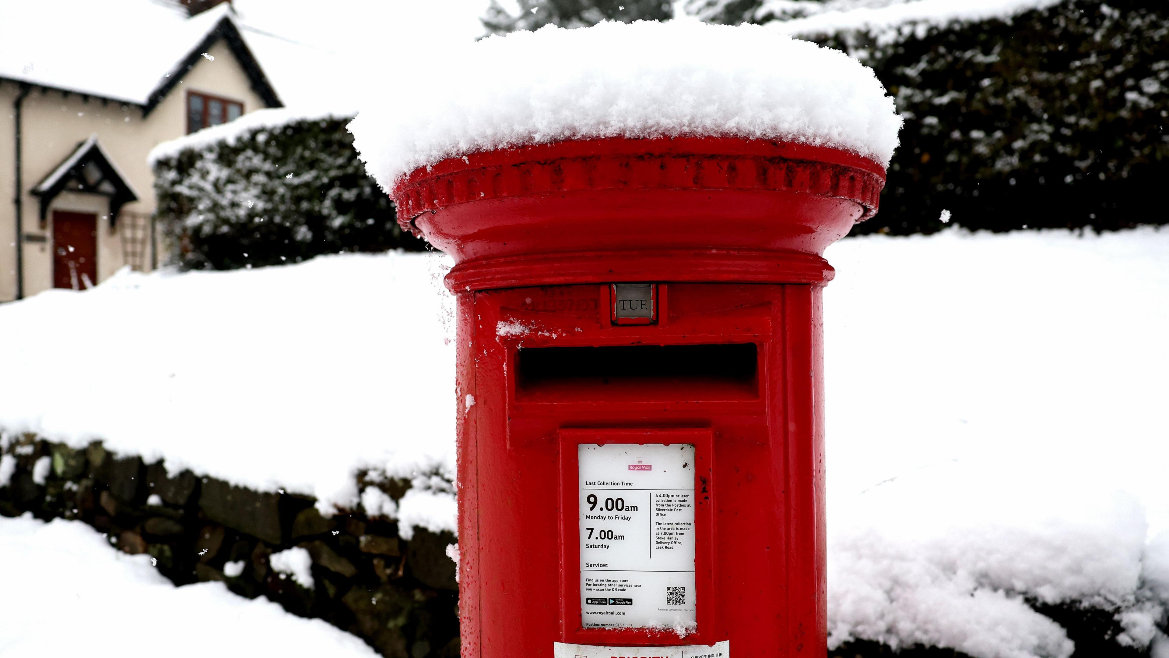 A red postbox with a mound of snow on top. Hedges and a house in the background are similarly covered in snow.