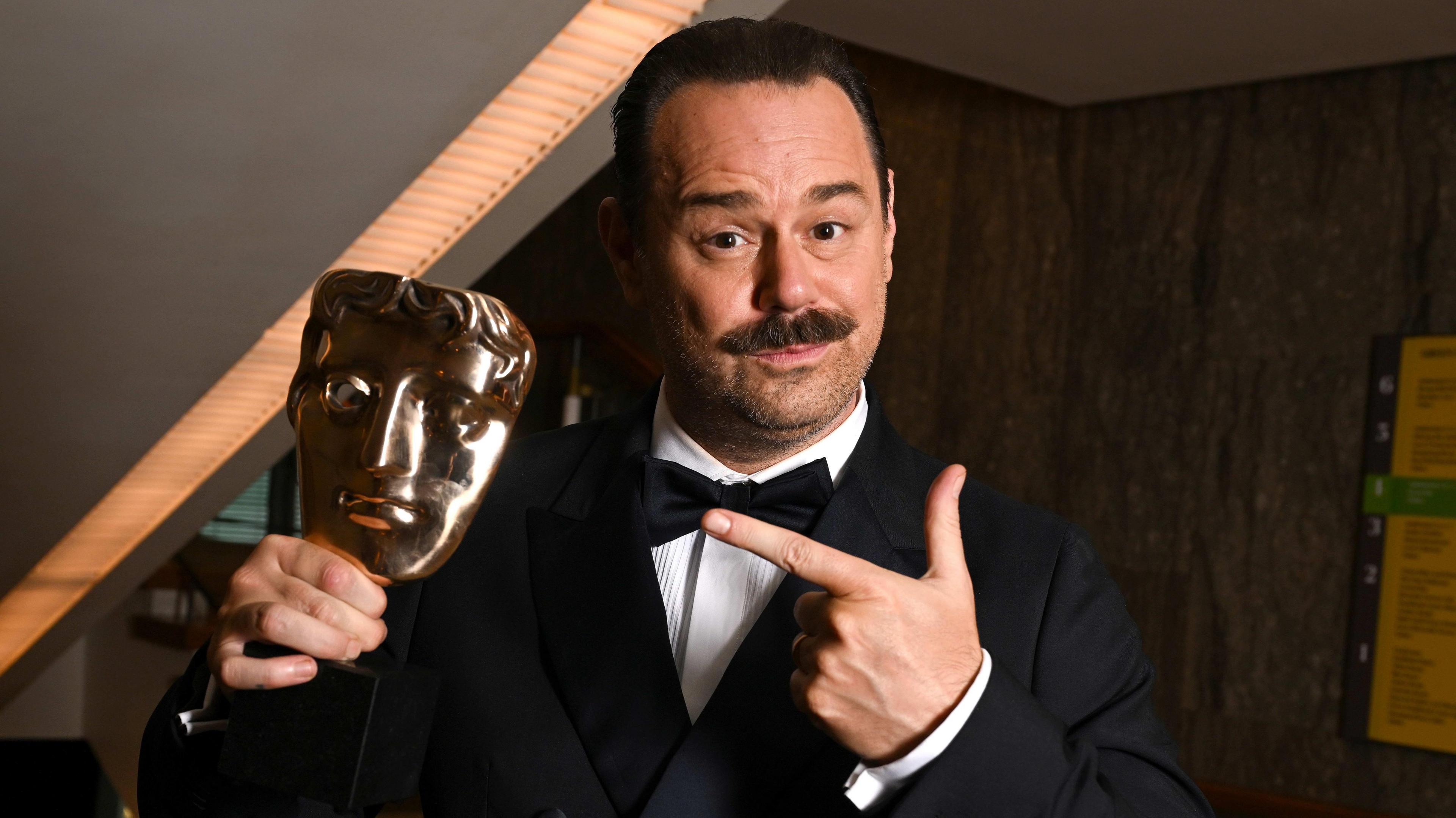 Danny Dyer in a black tux points to his Bafta which he is holding in his left hand. He has a moustache.