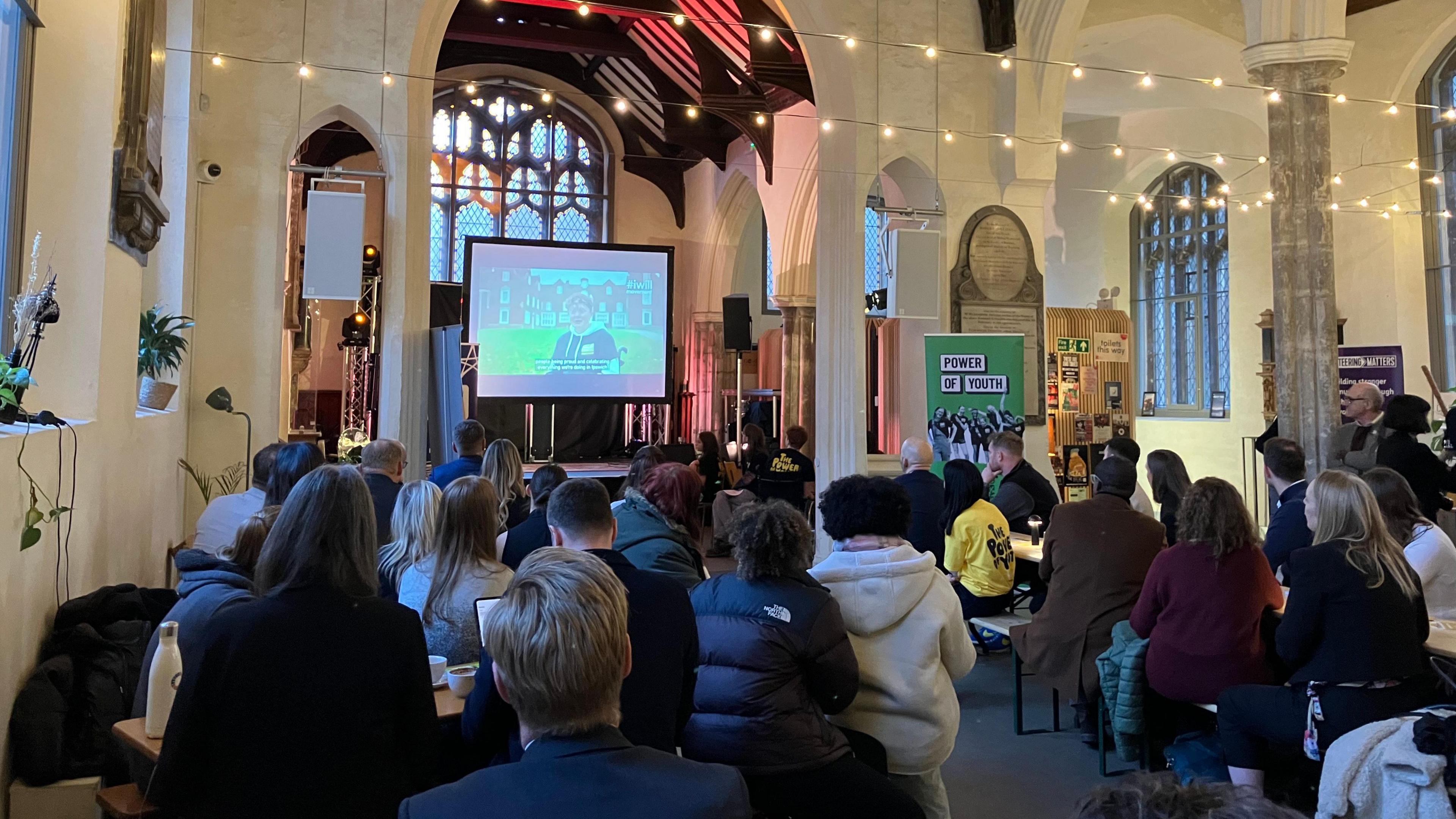 A crowd of people sat on benches and tables inside a church watch a large screen at the front