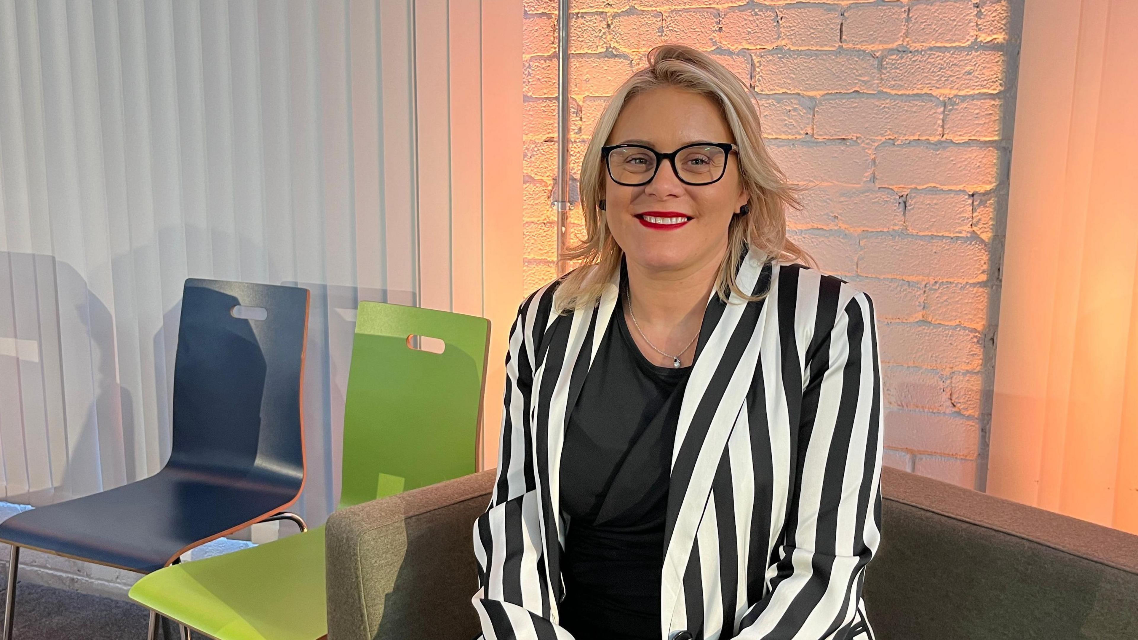 Councillor Emma Williamson sitting on a sofa and smiling at the camera. She is blond with blue eyes and wears glasses. She is wearing a black and white striped jacked over a black top.