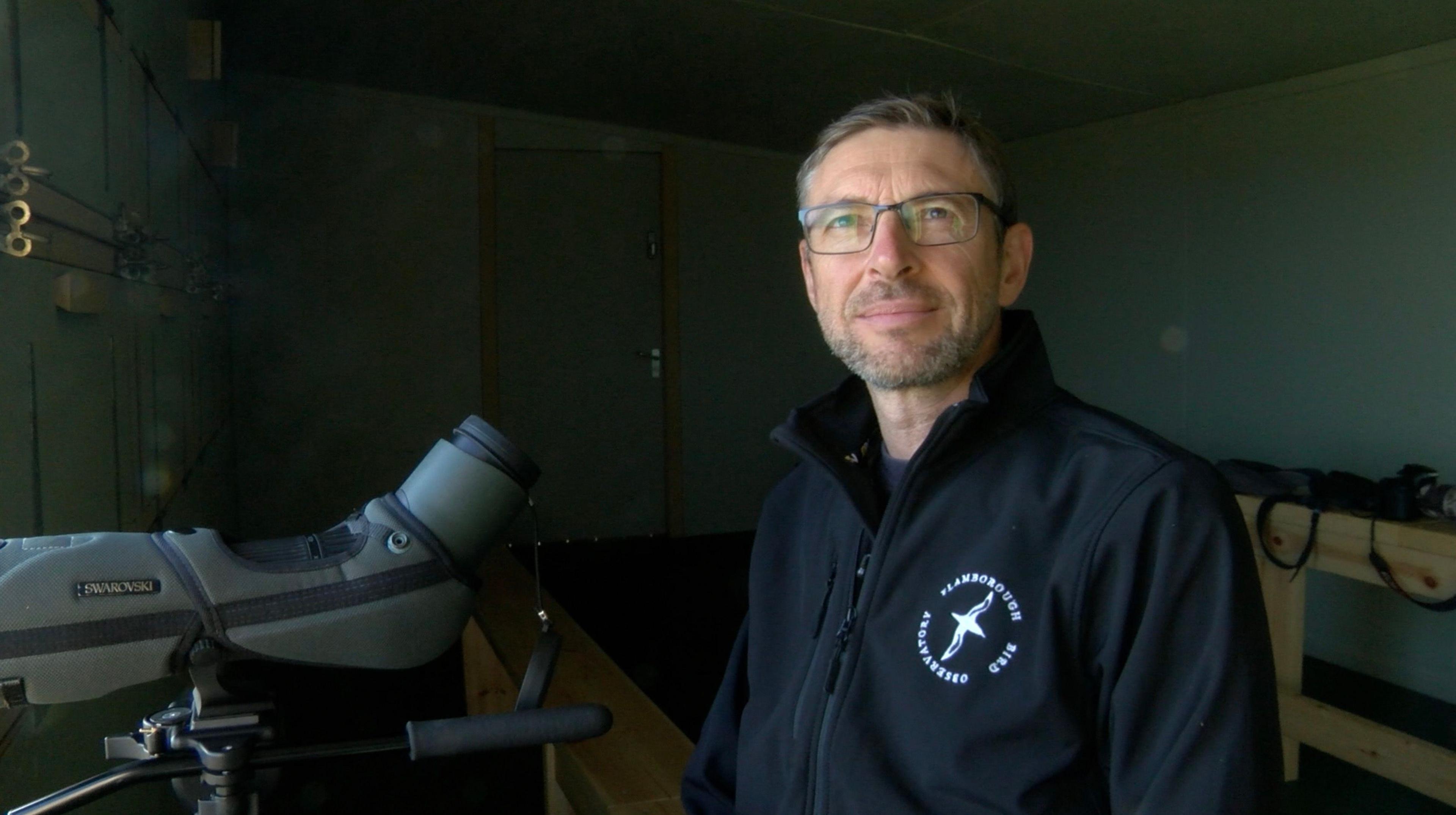 Mr Thomas is wearing a black fleece with a bird logo in white on the left hand side. He is standing in front of a telescope and is wearing glasses, he has grey stubble. 