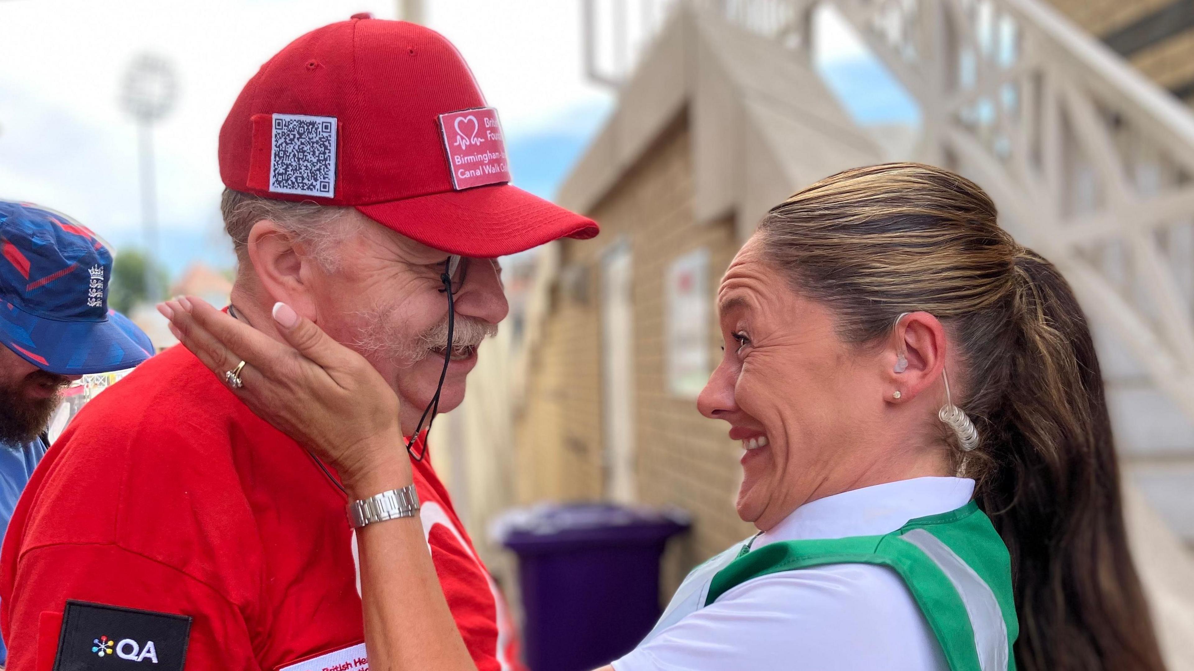 David Tomlinson and Hayley Spencer meet at Trent Bridge Cricket Ground. David is wearing a red t-shirt and cap. He has a moustache and wears a monocle. Hayley wears a green high-visibility vest over a white shirt. She is smiling and holding her hands up to David's face.