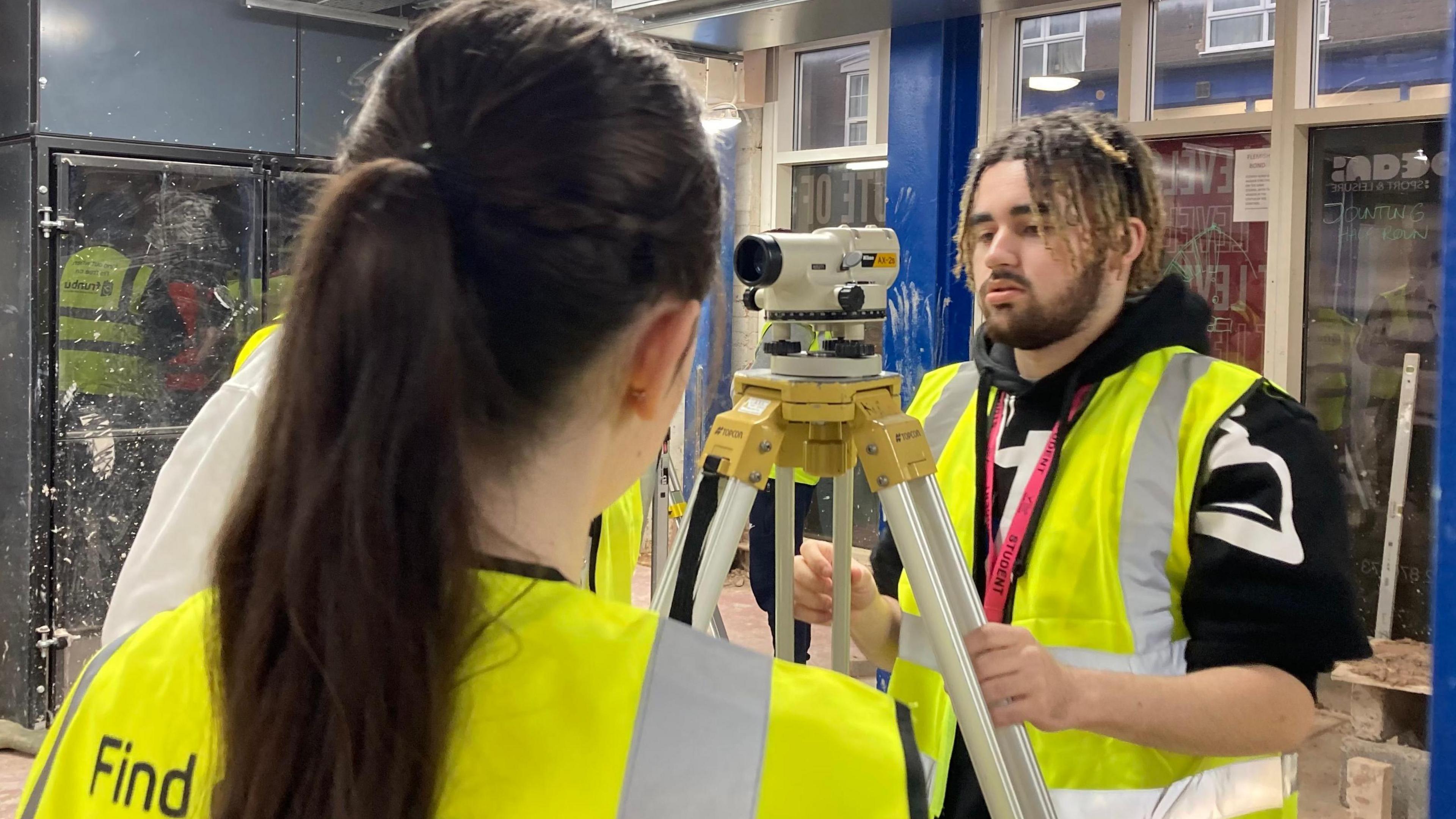 Two students wearing hi-vis jackets standing in a construction environment. A male to the right, with dreadlocks, is looking through a recording device, while a girl, with long brown hair in a ponytail, looks on with her back to the camera.