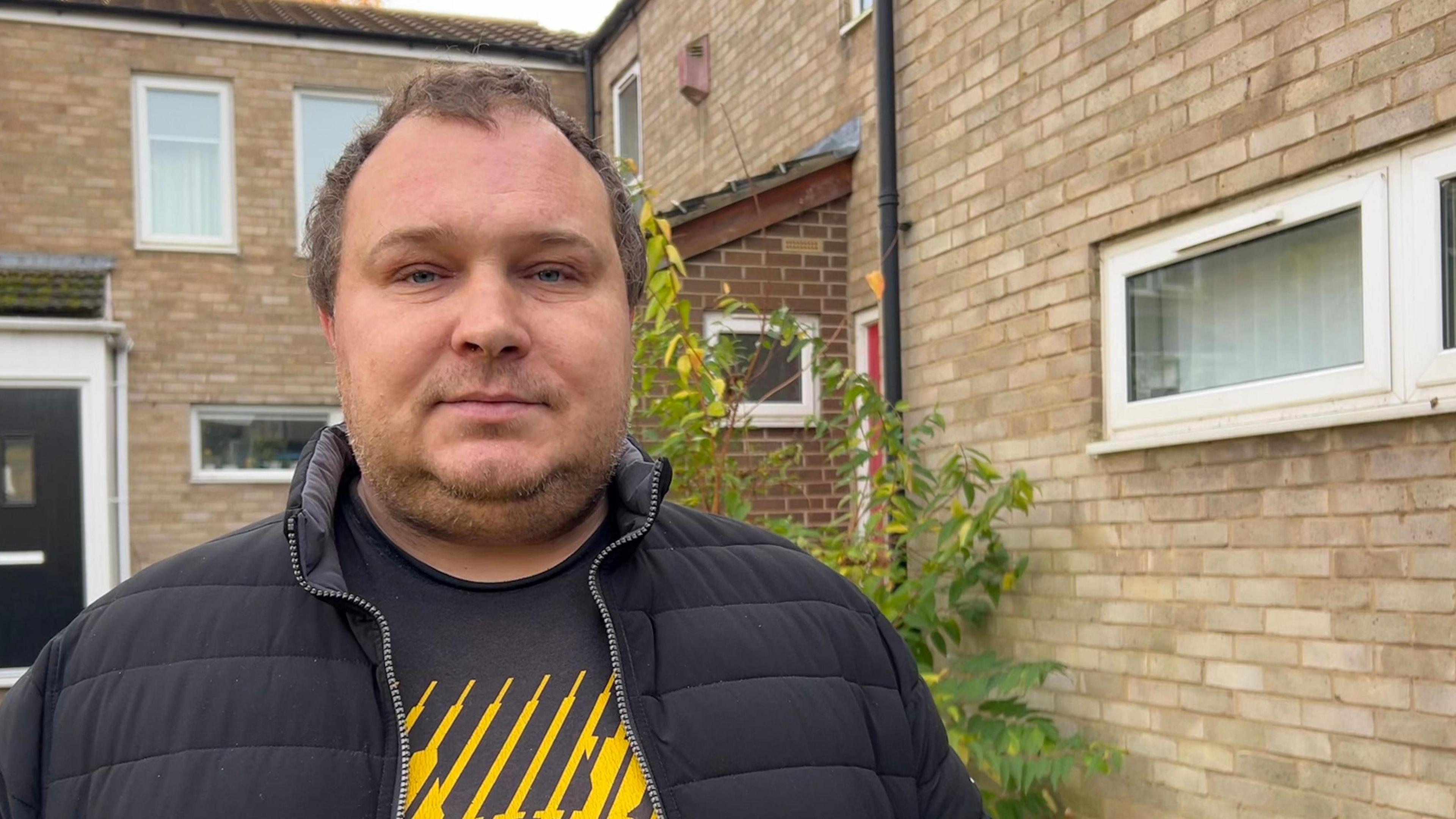 A man in a unzipped black coat. He is wearing a black t-shirt with a yellow striped design. He is standing outside a block of flats.