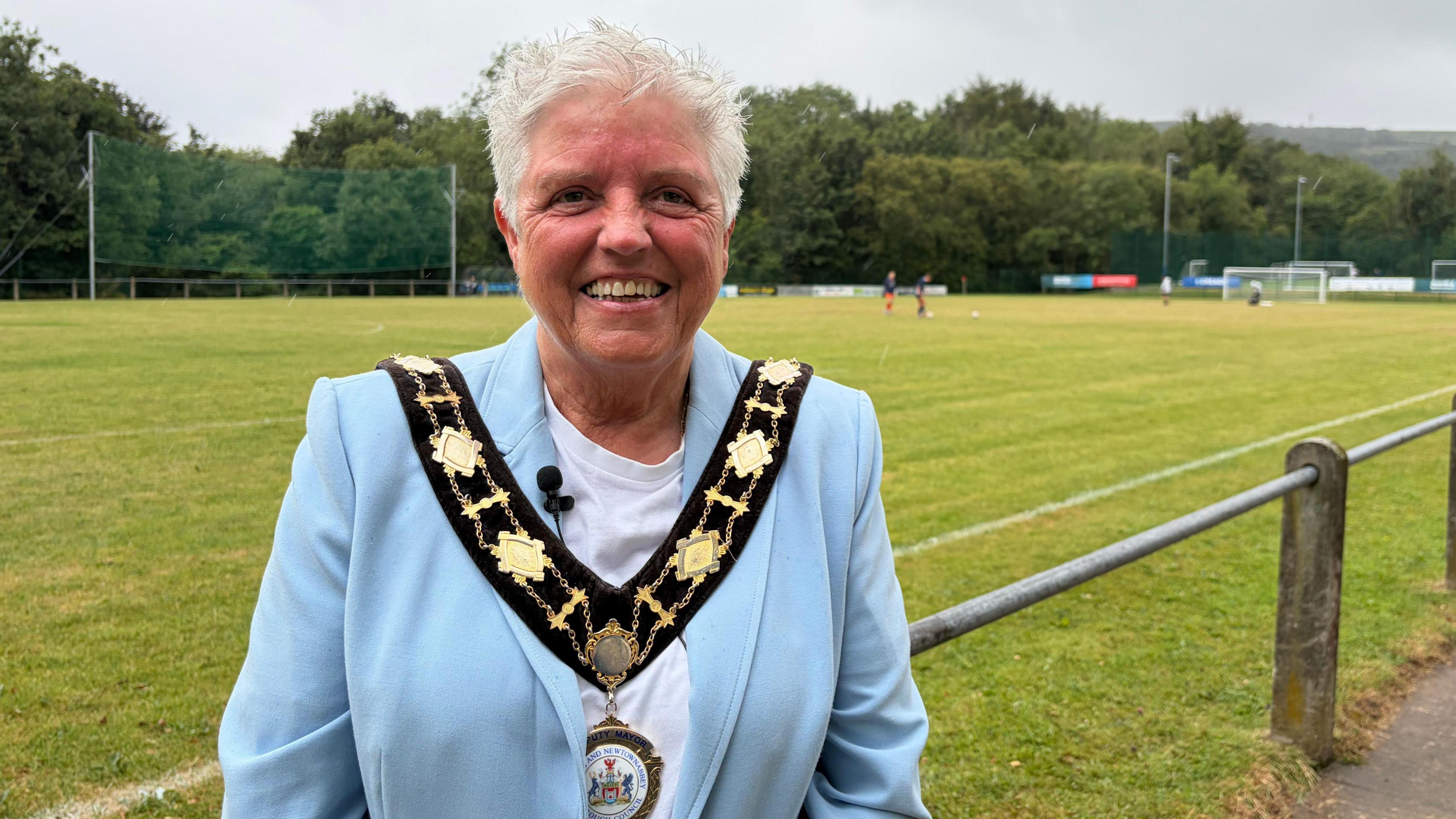 Alison Bennington stands in front of a football pitch. She has short grey hair and is wearing a blue suit jacket over a white T-shirt. Her deputy mayoral chain is over the top.