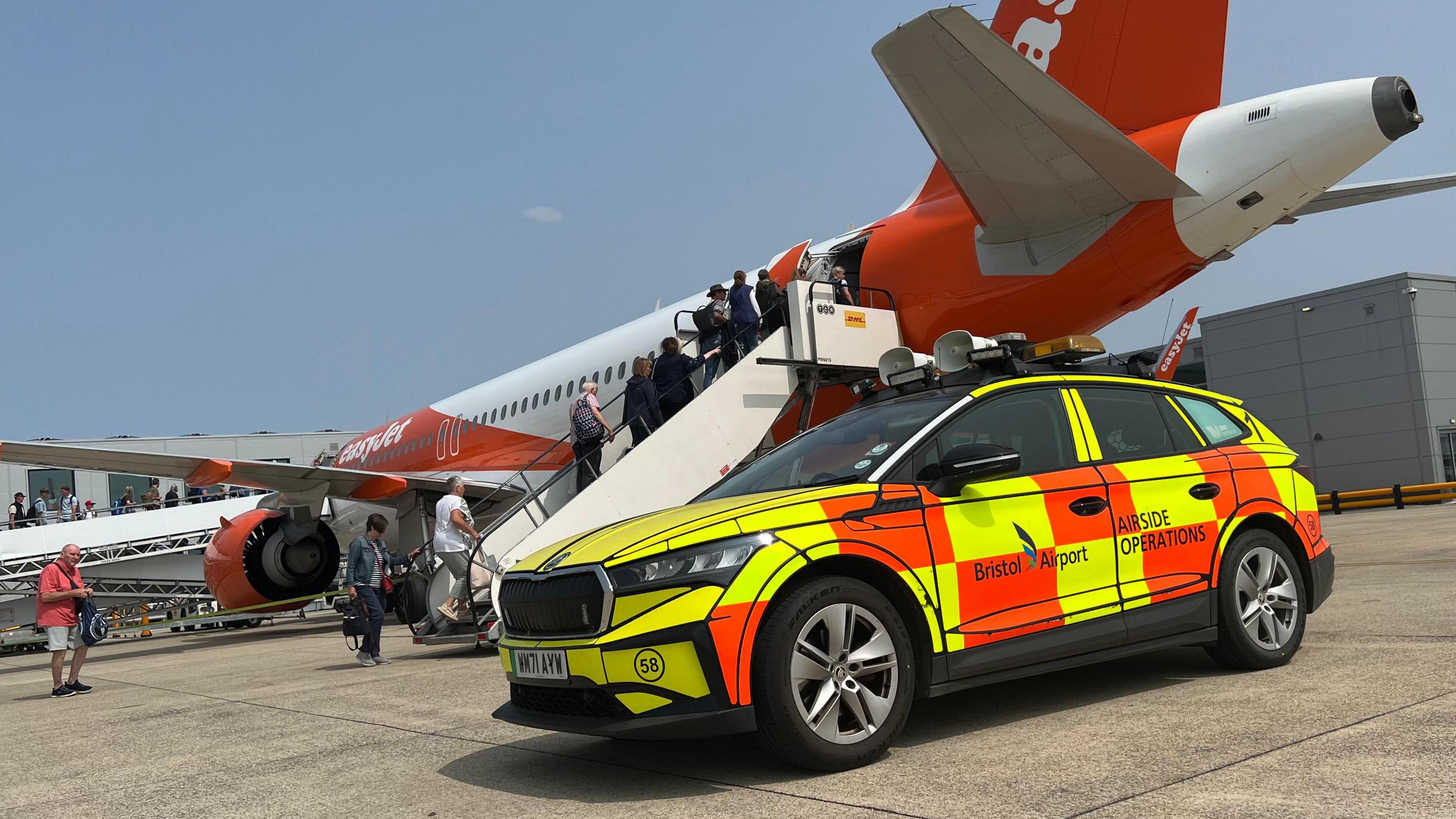 An electric car with yellow and orange cheques on an airport runway in front of a white and orange EasyJet plane. 