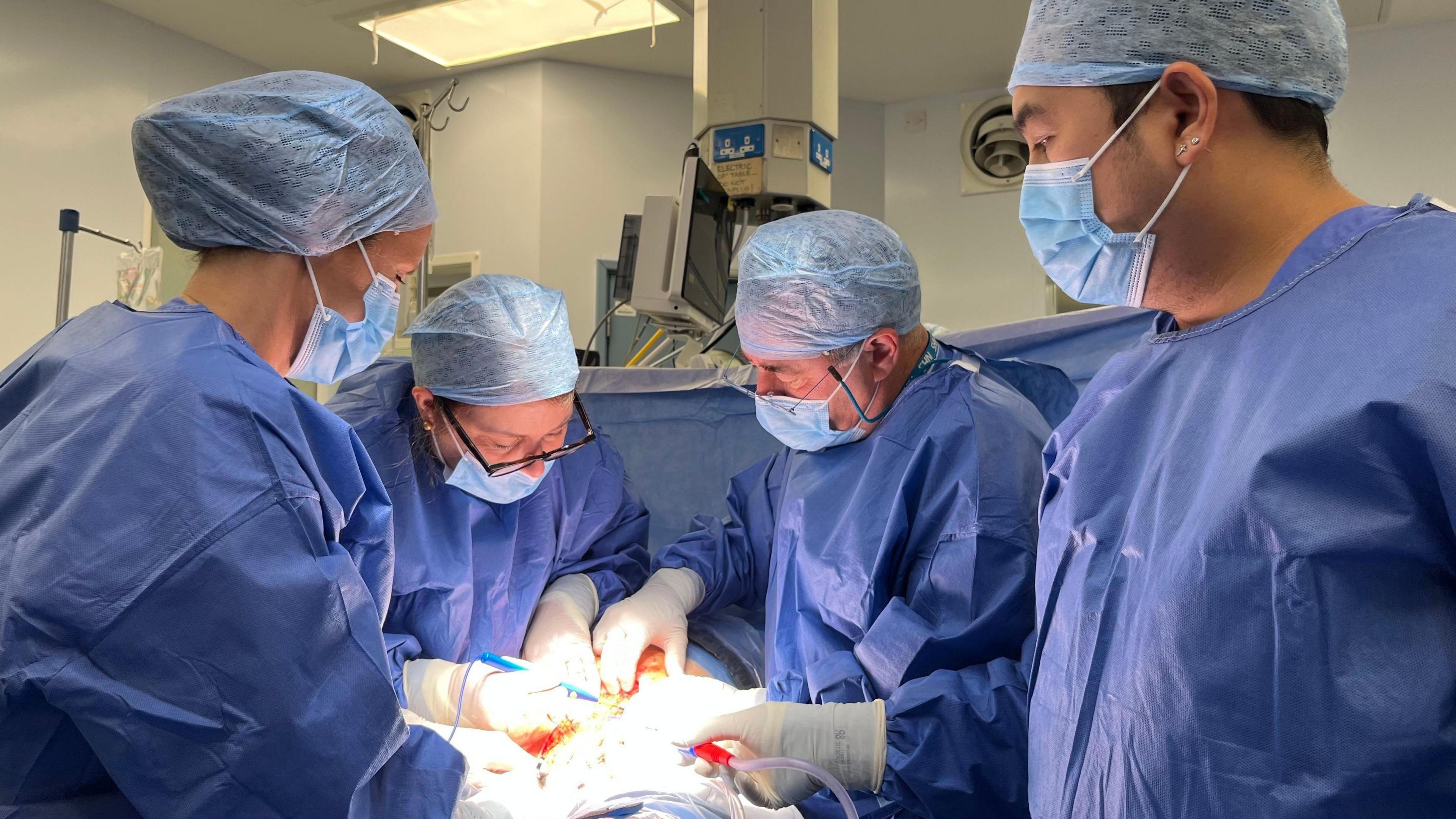 Katër kirurgë me uniforma mjekësore duke kryer operacionin e transplantimit të mitrës