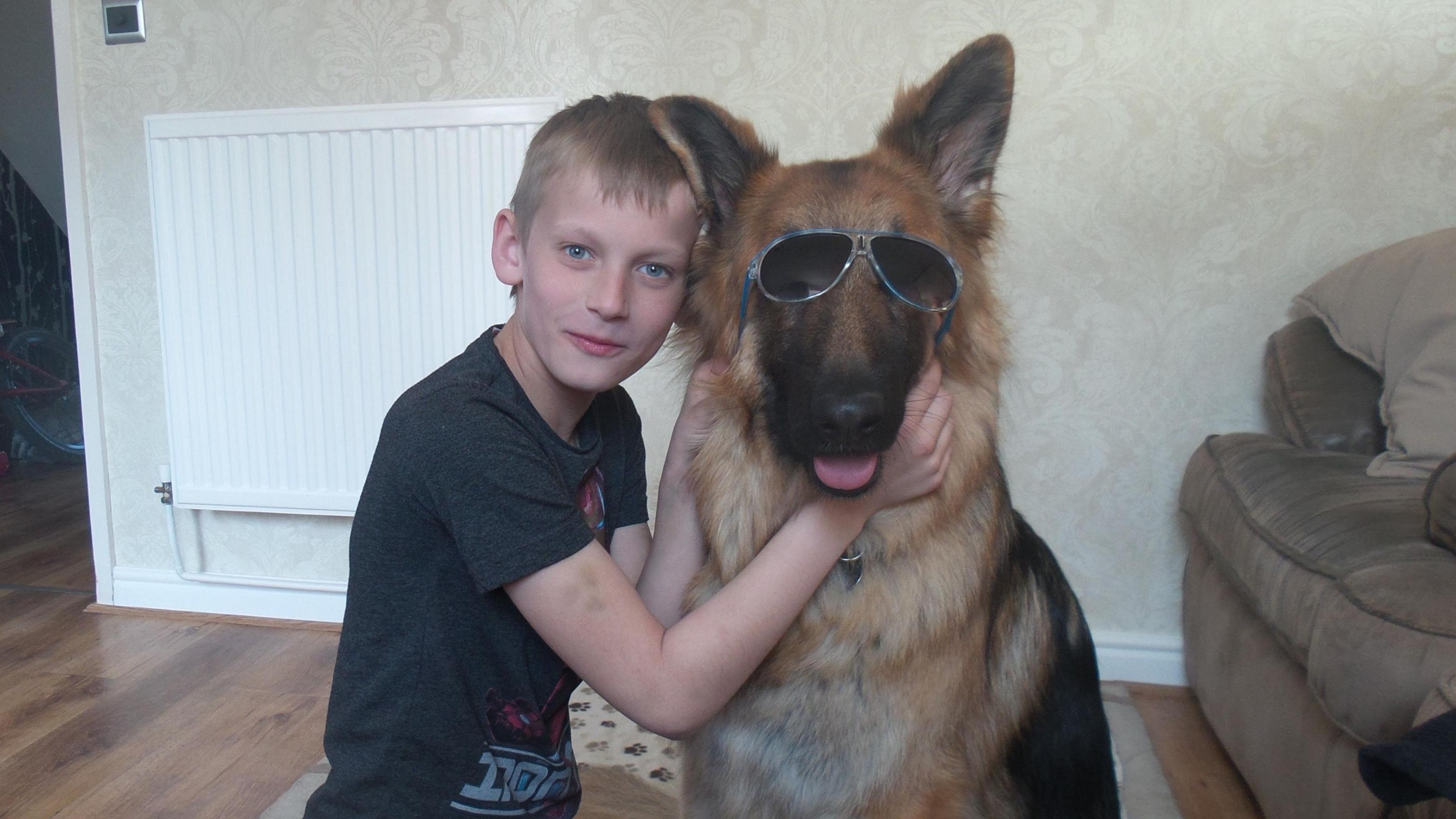 A childhood picture of Ben Moncrieff kneeling on the floor of his living room beside his childhood dog, Barney. It is a large brown and black German Shephard, and Ben is holding a pair of blue sunglasses over his eyes to pose for a photo. In the background there is a patterned cream wallpaper, wooden floors, a white radiator and a grey sofa. 