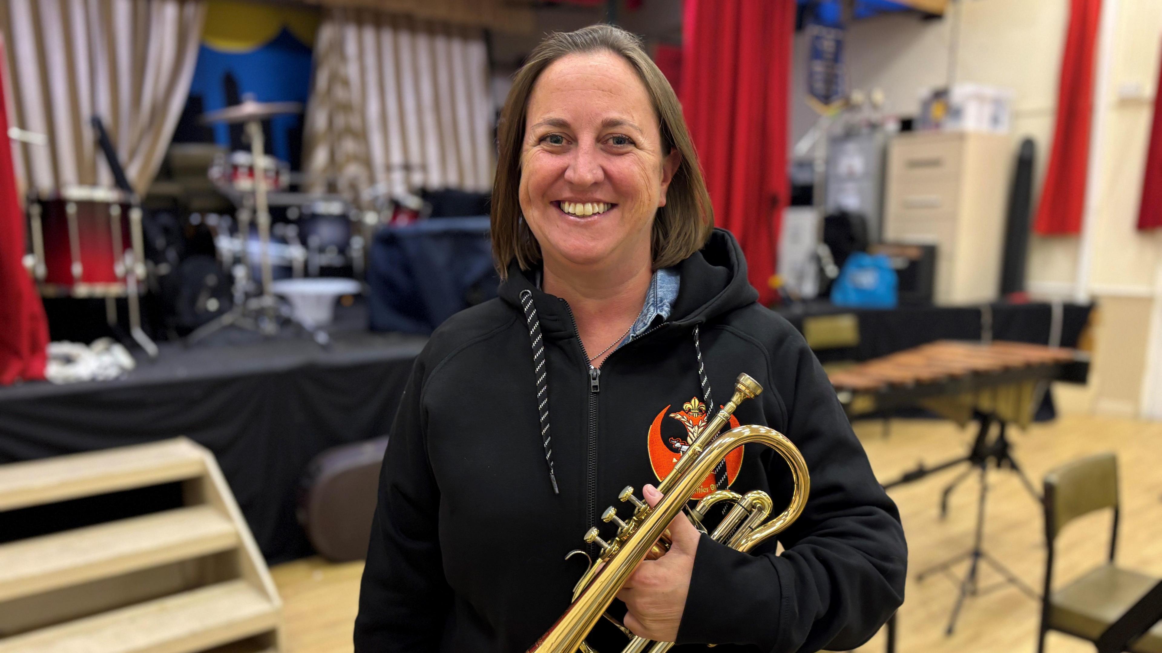 Vanessa Blanchet is wearing a black zip-up hoodie and is holding a gold-coloured cornet. She is in a room with a stage behind her. There are drums on the stage.