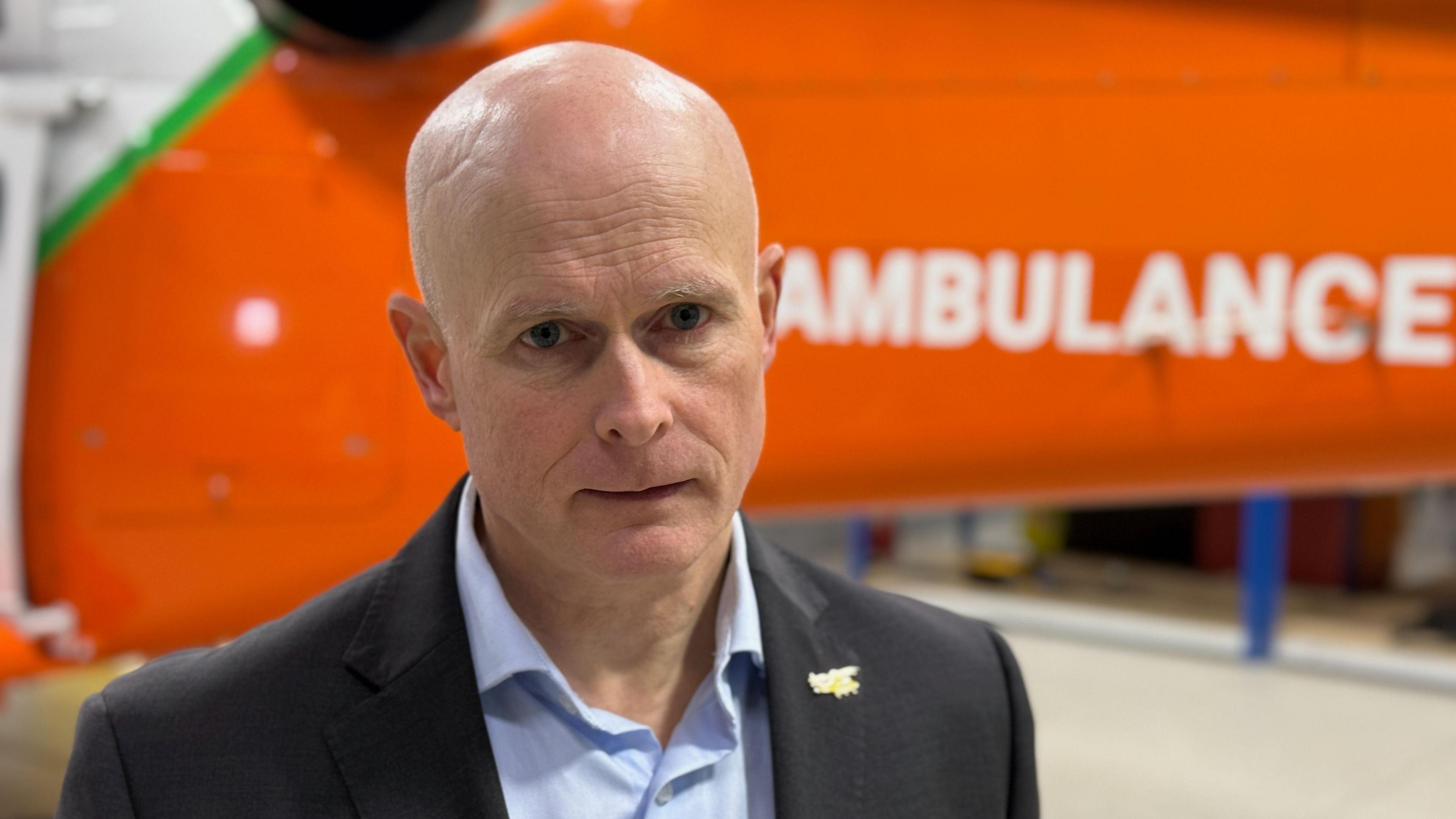 Dr Phil Hyde poses in front of an orange helicopter which says 'ambulance' on the side. He is bald, and wearing a light blue shirt underneath a dark blue suit jacket