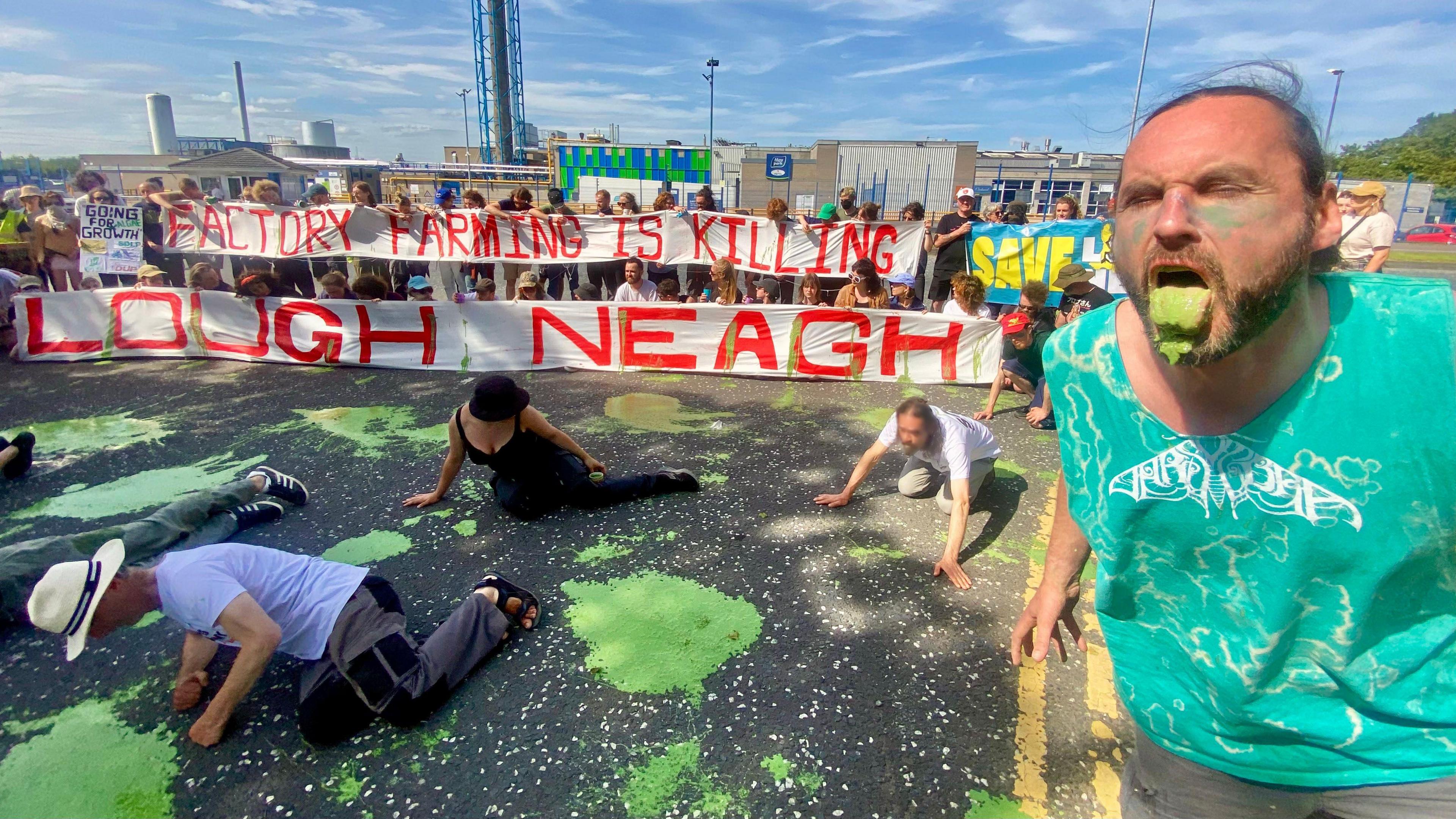 A man with his mouth open and tongue out with green sludge in his mouth. Other people lie on the ground next to puddles of the sludge while in the background people are holding up a large banner that reads "Factory farming is killing Lough Neagh".