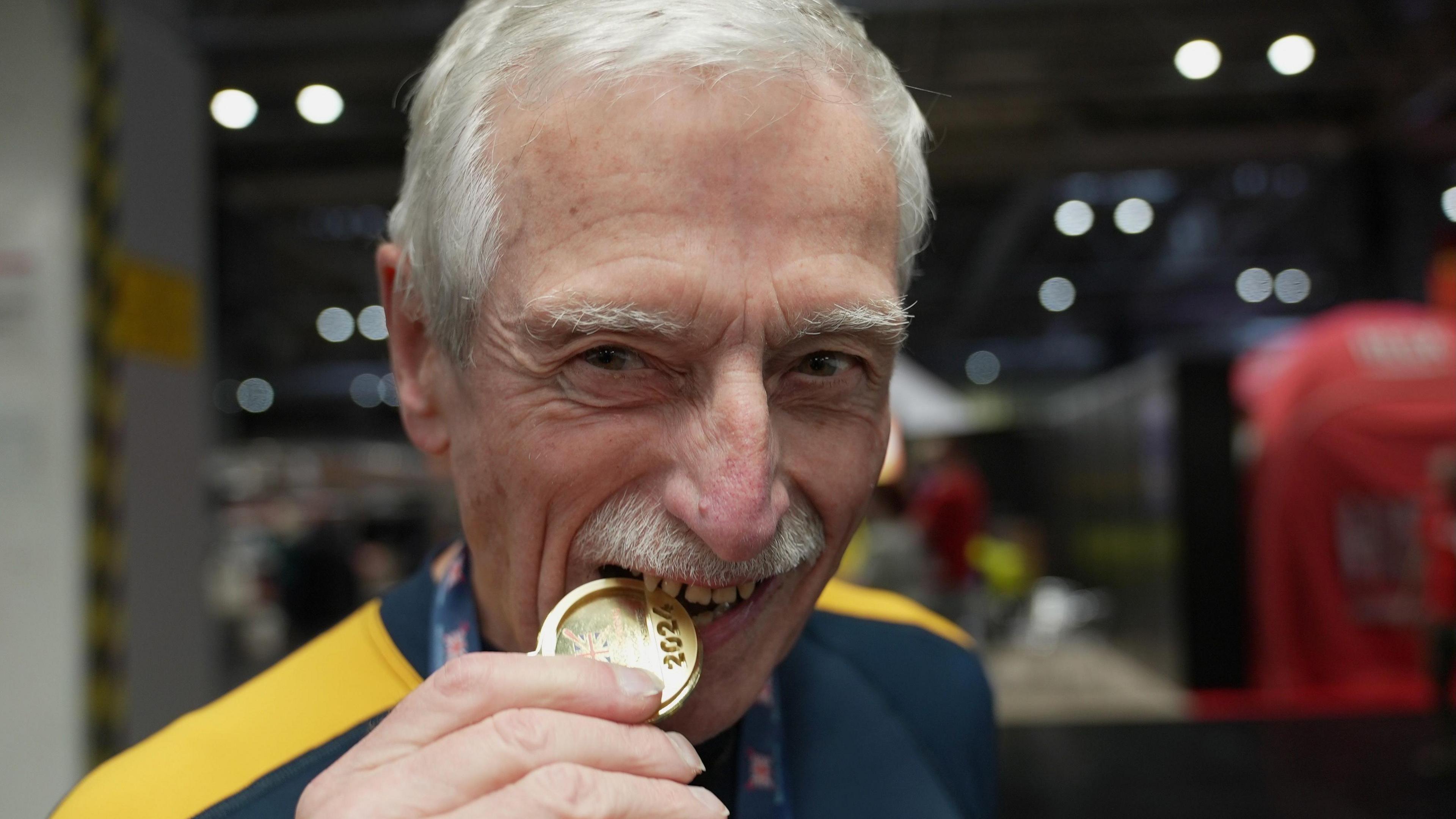 Roger Stainforth has grey hair and a moustache. He is looking directly at the camera and is biting his gold medal.