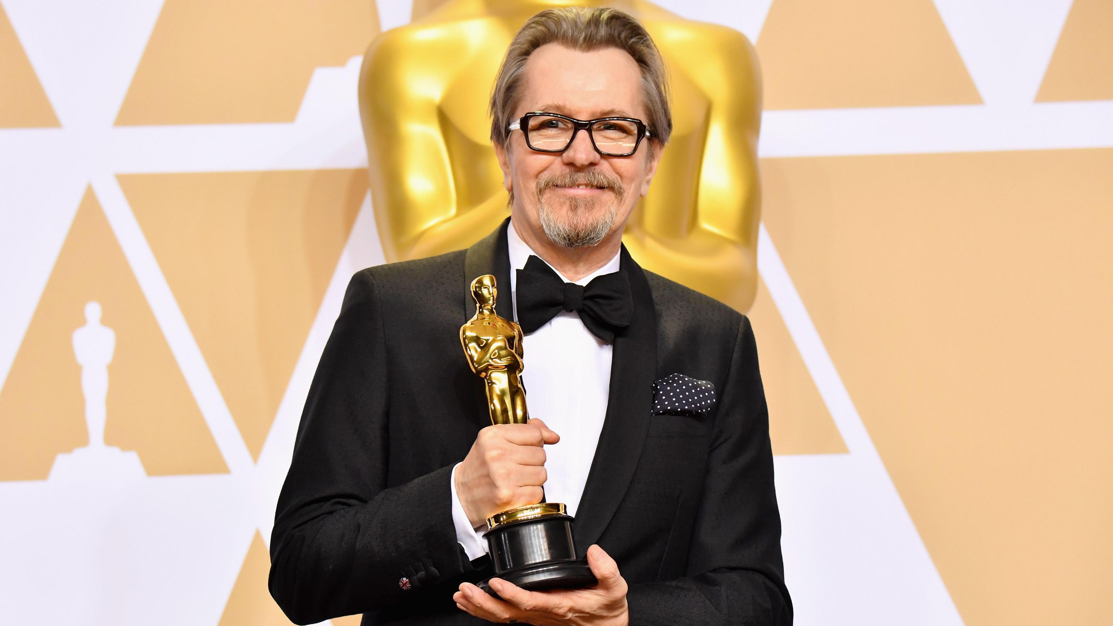 Gary Oldman is wearing a black and white tuxedo with a black bow tie. He is holding his Academy Award trophy which is a golden statue of a man. 