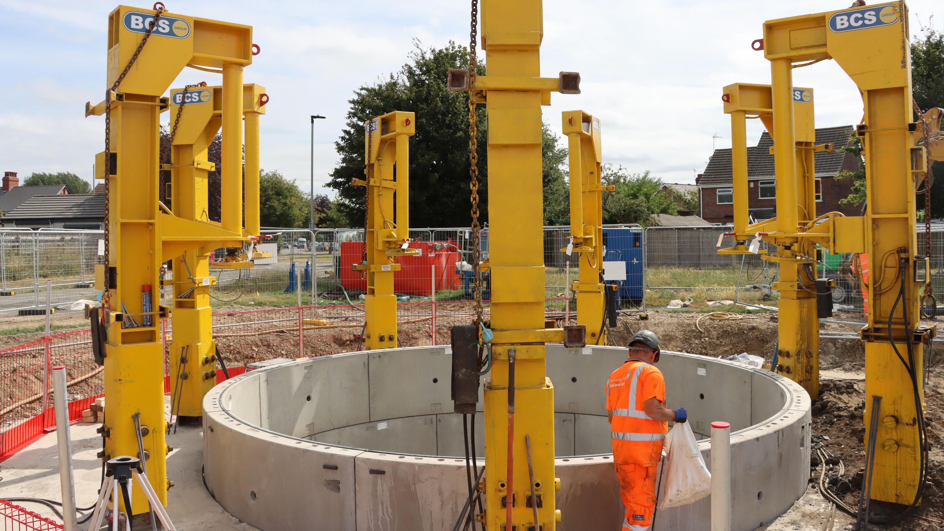 The new tank is outside and has been made of light grey concrete blocks, in a circle shape. Around it are large yellow metal devices. A worker in high visibility uniform is stood next to the tank and holds a white plastic bag. 