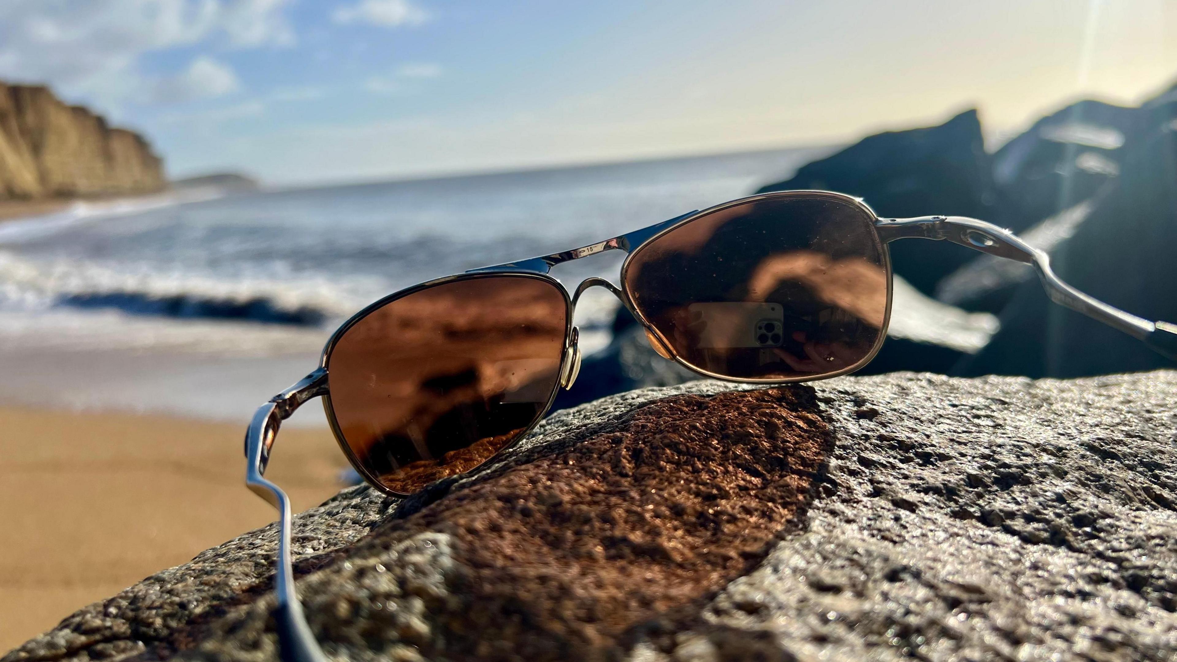 A pair of sunglasses are perched on a rock. The sunglasses are in focus. Behind you can see a beach on a sunny day with waves lapping on the sand. There are cliffs in the distance. 