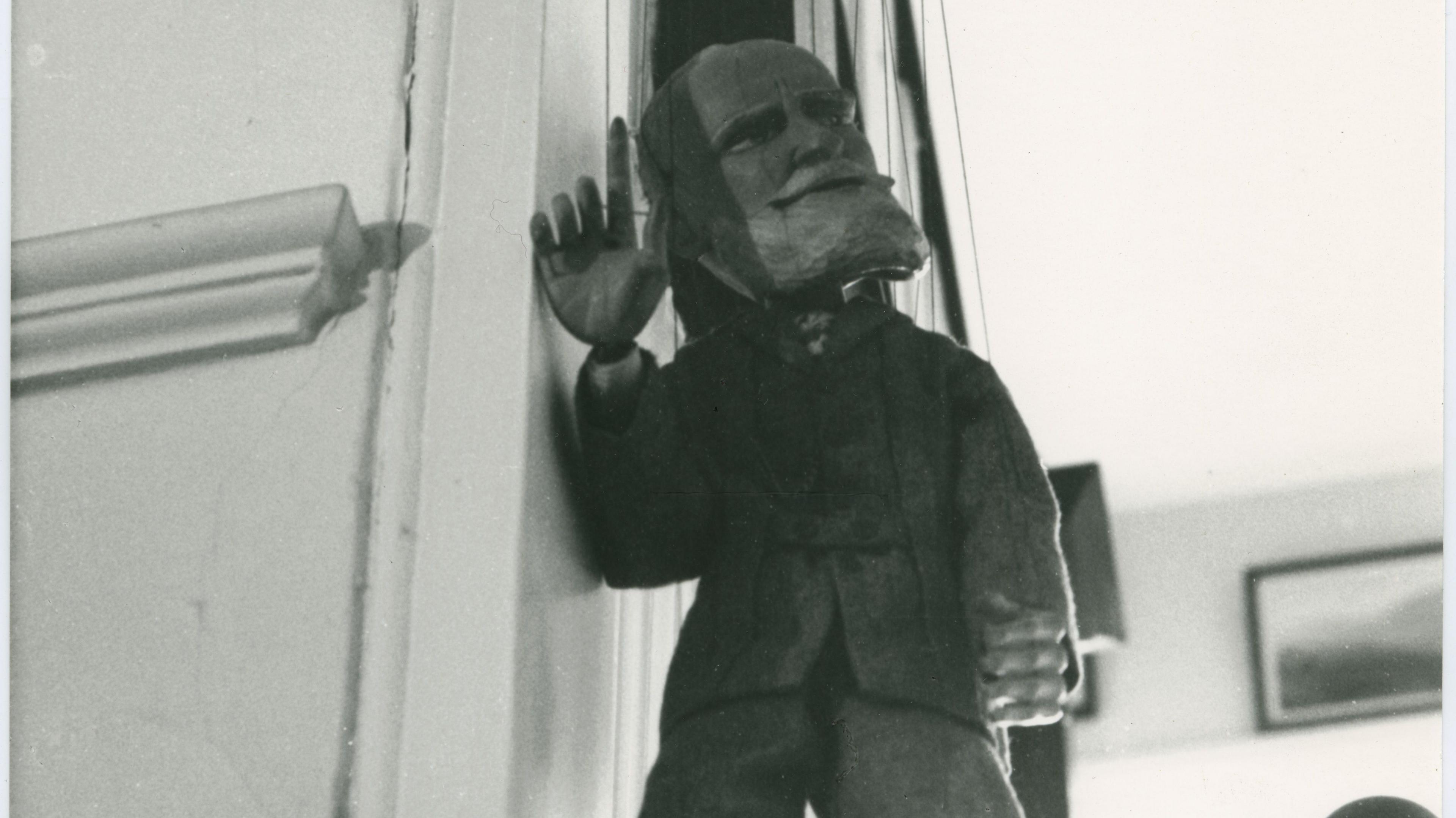 Black and white photograph of a puppet dressed in a suit with one hand raised.