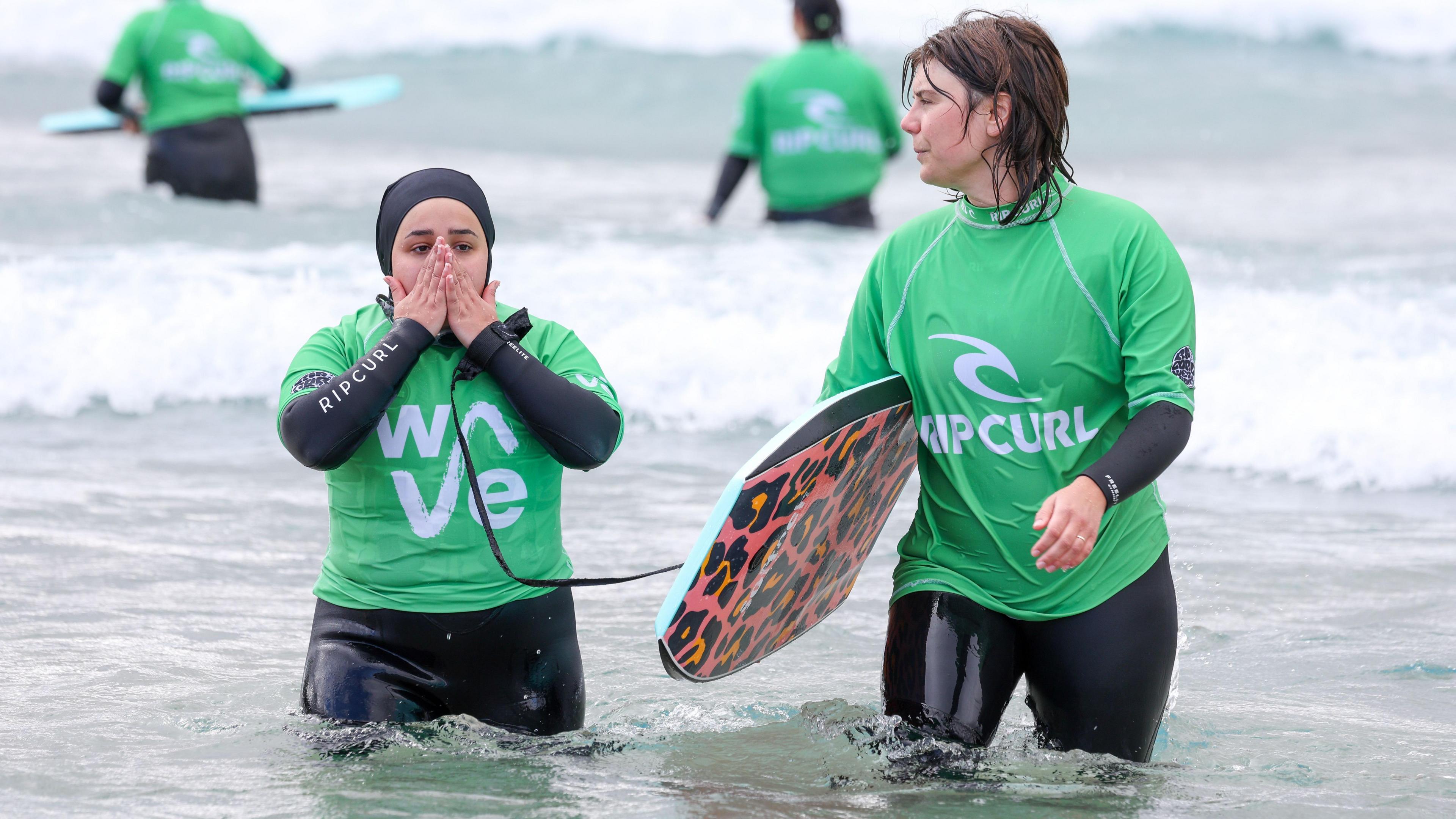 A participant wading through the water on the left wearing a black wetsuit, green rash vest, and waterproof hijab. She is holding her hands up to her face while a volunteer walks beside her, carrying a bodyboard with leopard print on the bottom. The bodyboard leash is attached to the participant's wrist.