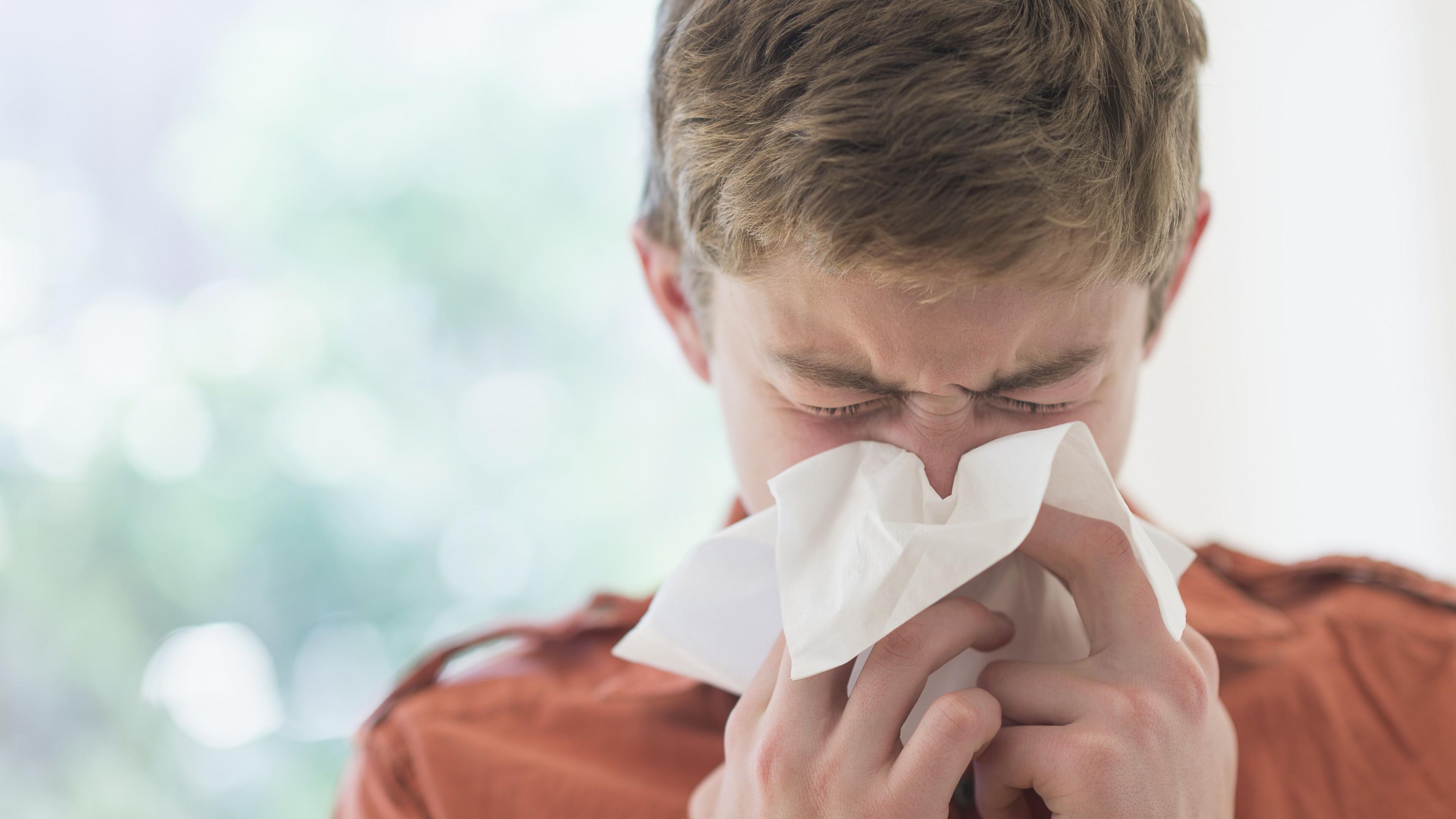 A boy in an orange shirt sneezes into a tissue with his eyes closed.