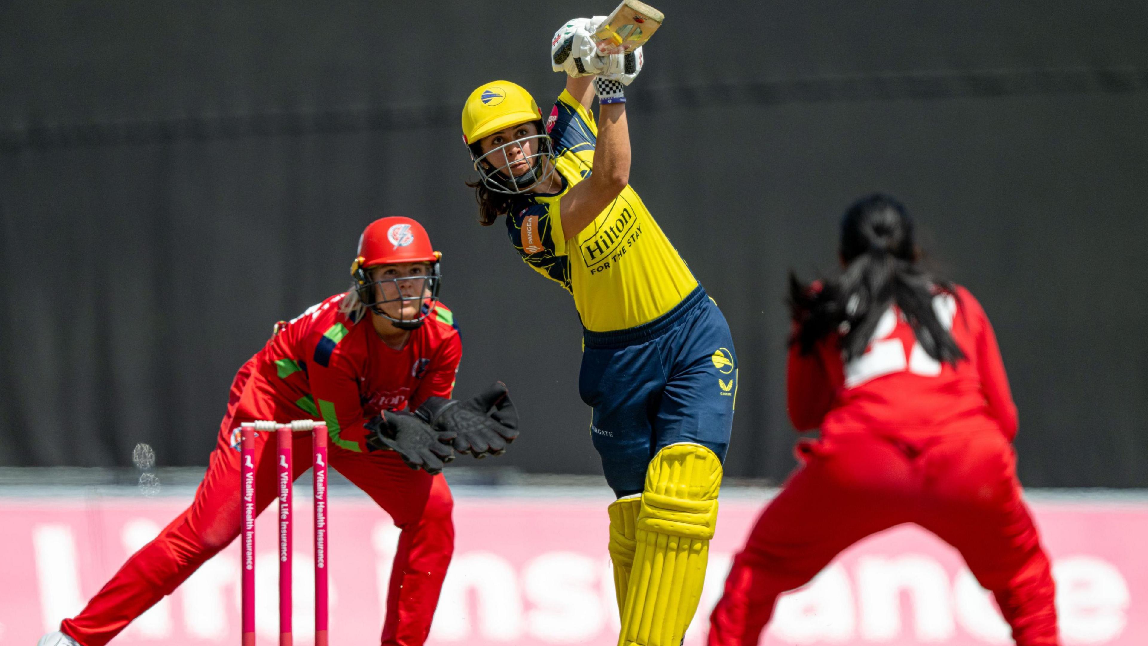 Maia Bouchier in yellow shirt, helmet and pads, and navy trousers, drives with Lancashire wicket and bowler, in all red, also in shot