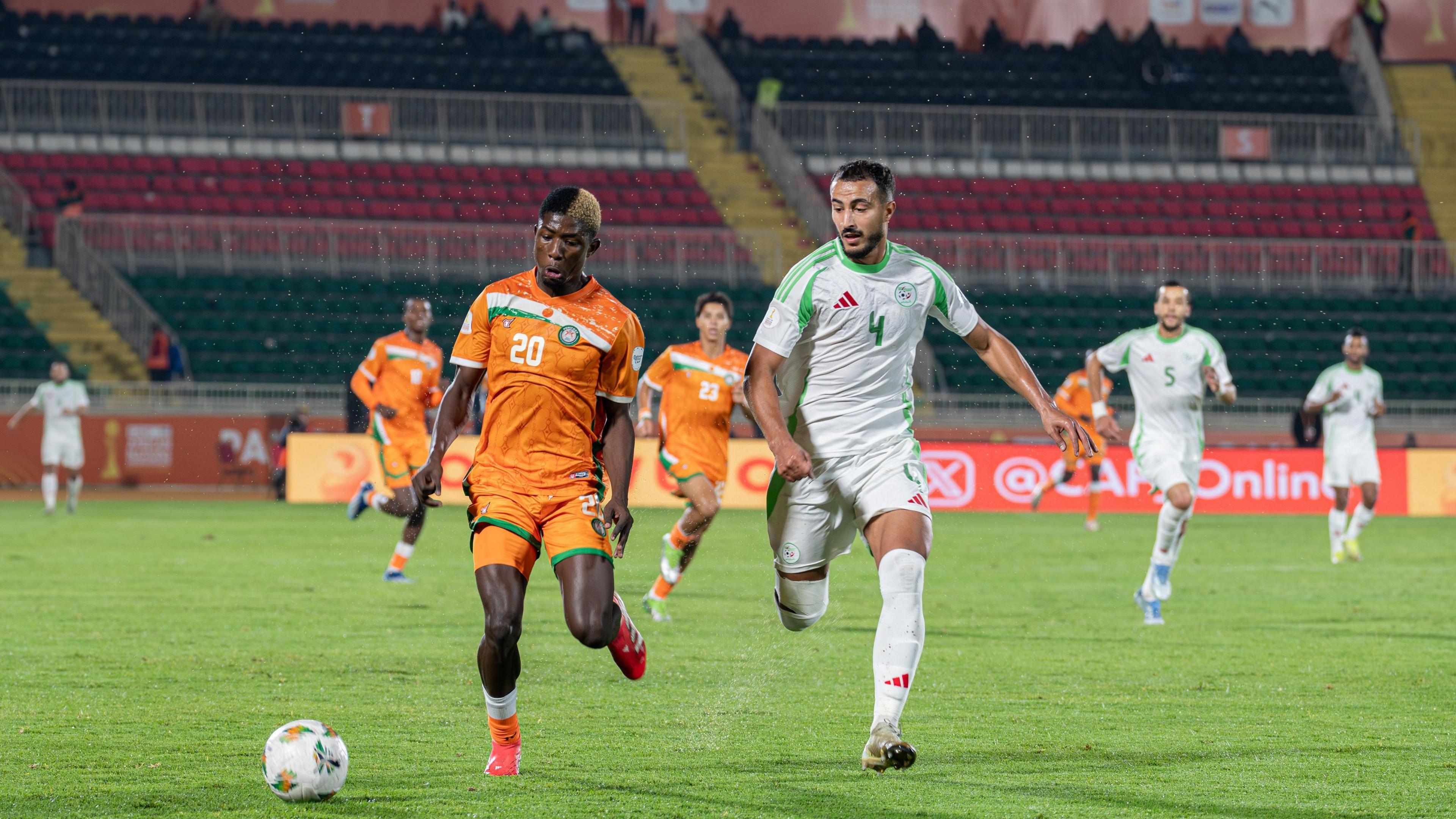 Adem Alilet of Algeria, wearing an all-white strip with green trim, chases Abdoul Latif Djibril Goumey Diori of Niger, who wears and all-orange shirt with white and green trim, during an African Nations Championship group match. Largely empty stands are seen in the background