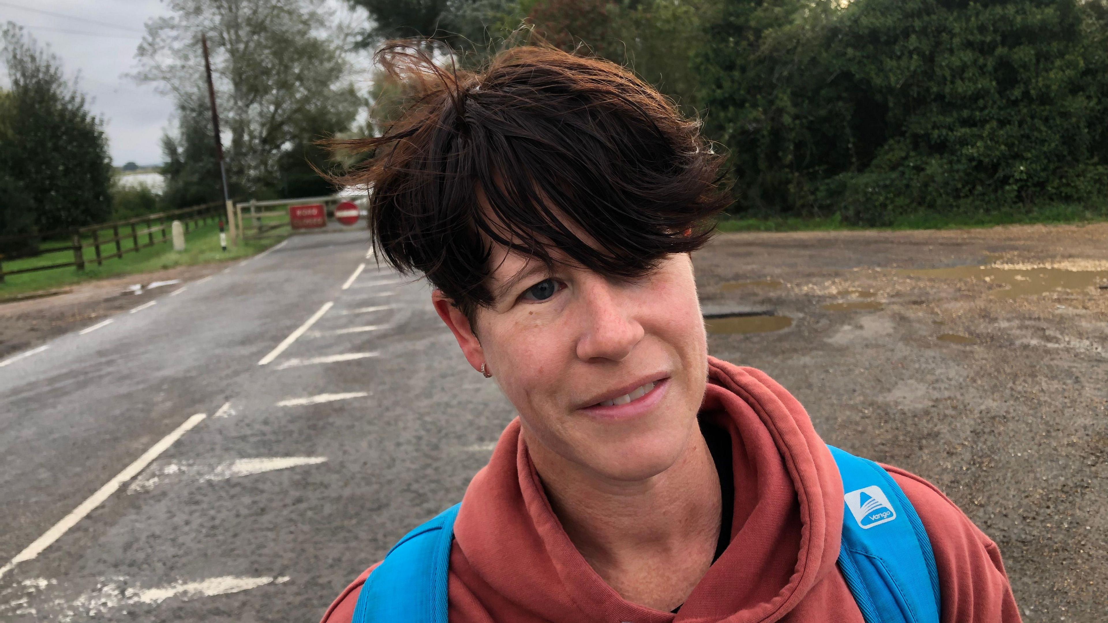 Lee-Anne Bower has a burnt orange coloured hoodie top, with blue rucksack straps over her shoulders. She has short dark hair which sweeps over her forehead. She is standing in front of the flooded road.