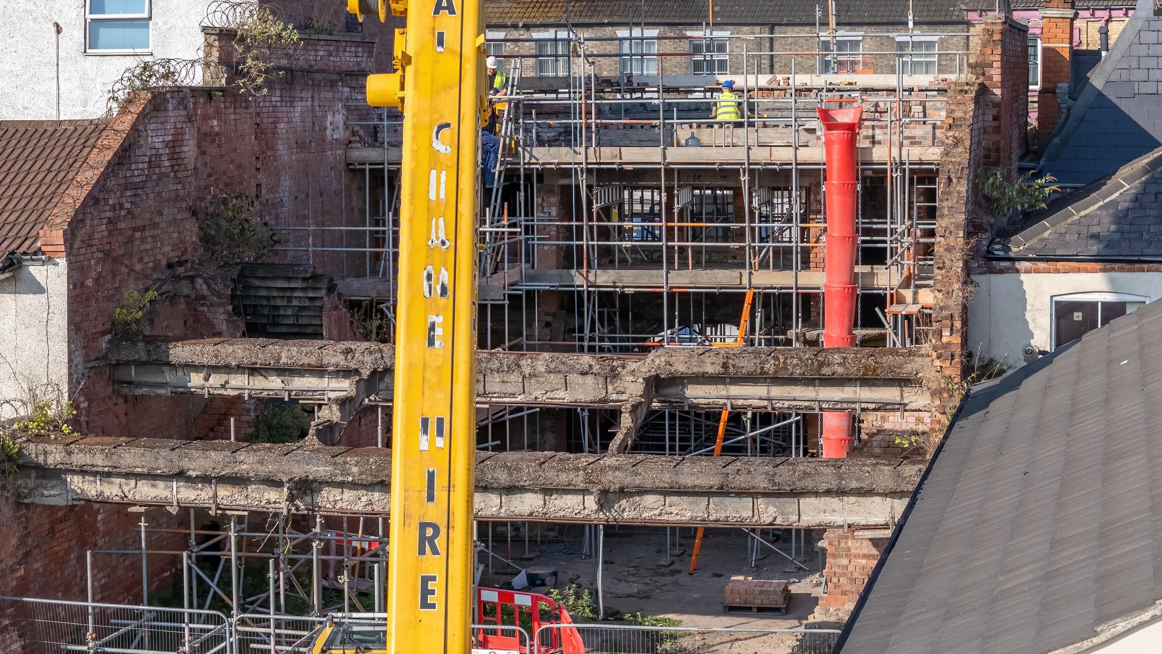 A view of the cinema from the rear, including two concrete beams which survived the blast. The building is overgrown with plants and covered in scaffolding. A yellow crane can be seen in the foreground.