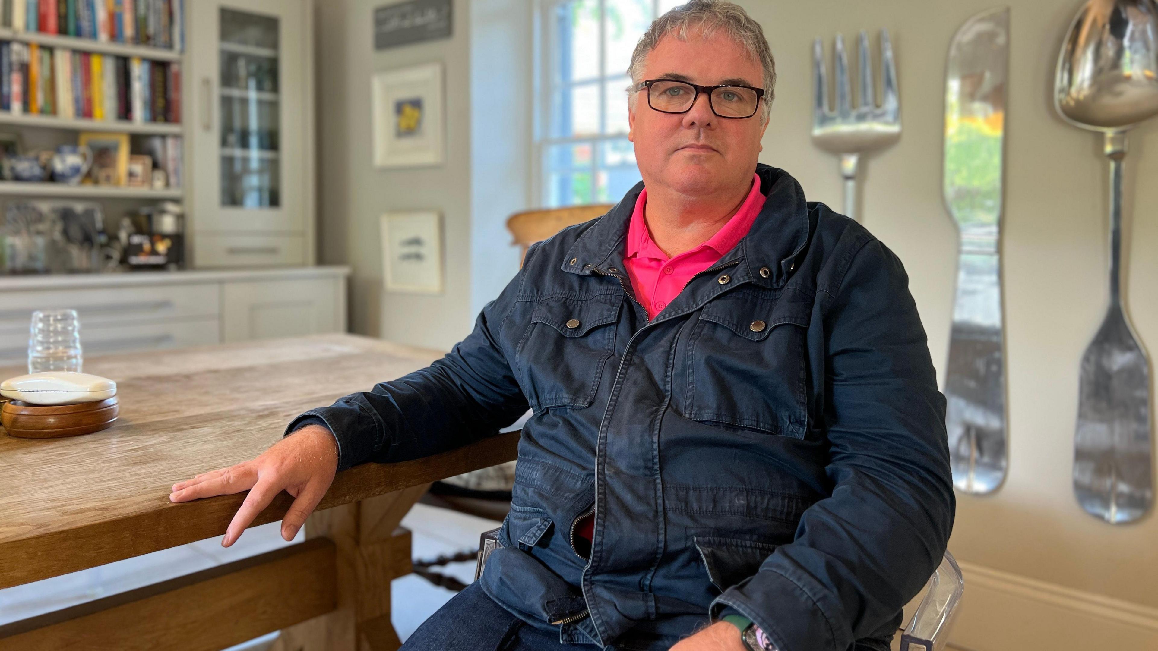 Stephen sits in a kitchen looking at the camera. His left hand is on the kitchen table which is next to him. He's wearing a dark blue denim jacket with a pink, collared t-shire under it. Behind him is a cabinet with books to his left and to his right there are large decorative cultarly hanging from the wall.