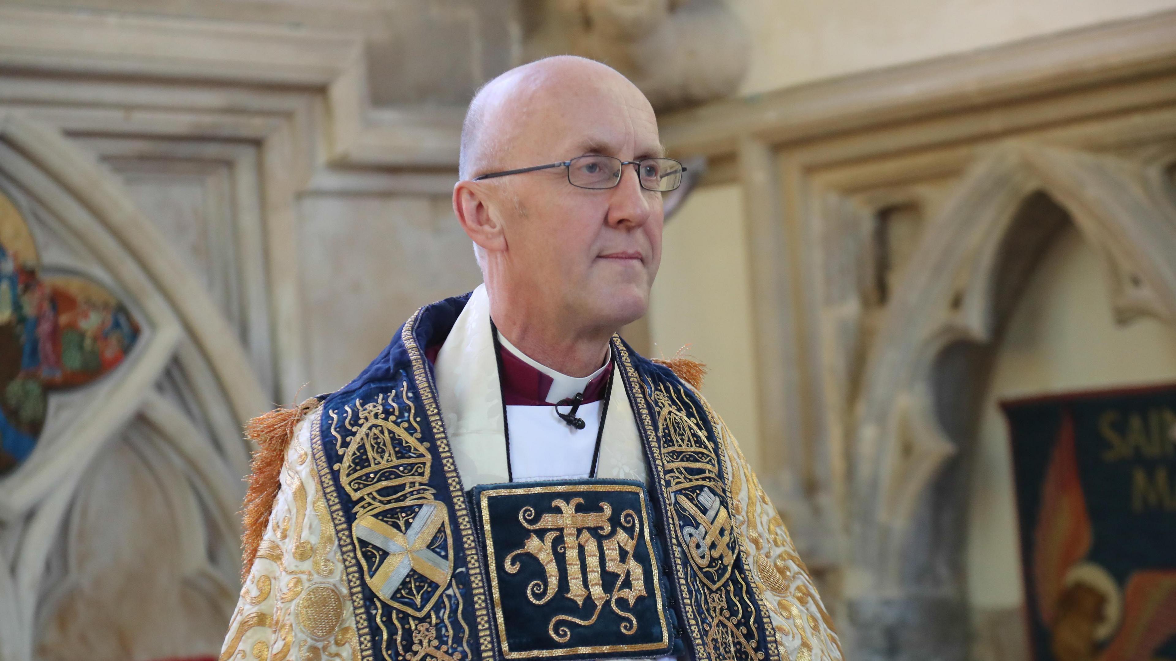 Bishop Michael Beasley is pictured in his ceremonial robes looking off to the right. He is standing in a church, with arches behind him. 