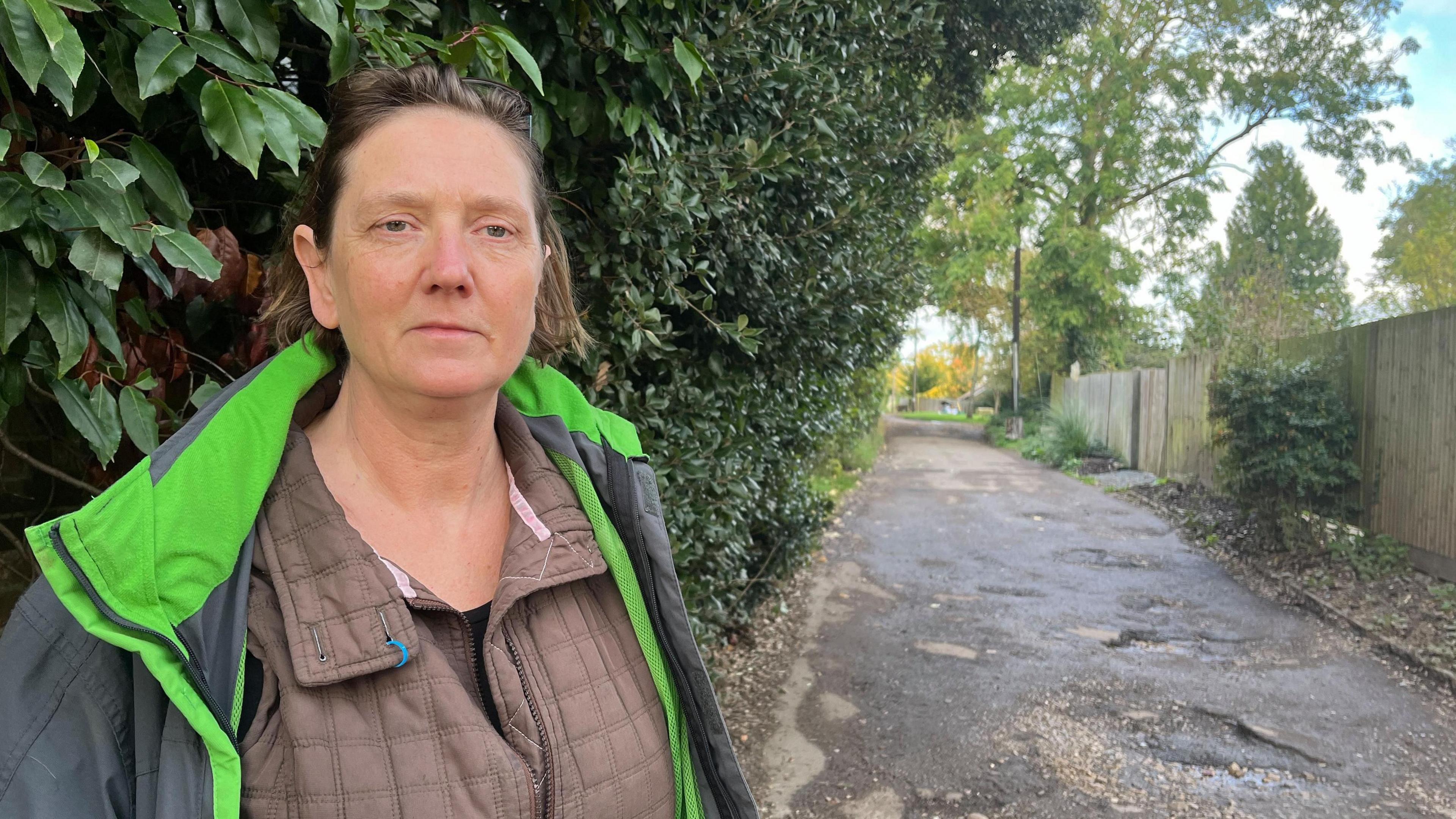 Sarah Wright standing by a pothole-ridden road. She is wearing a grey-green anorak over a padded brown jacket. She is looking towards, but not directly at the camera. Trees and a wooden fence can be seen behind her.