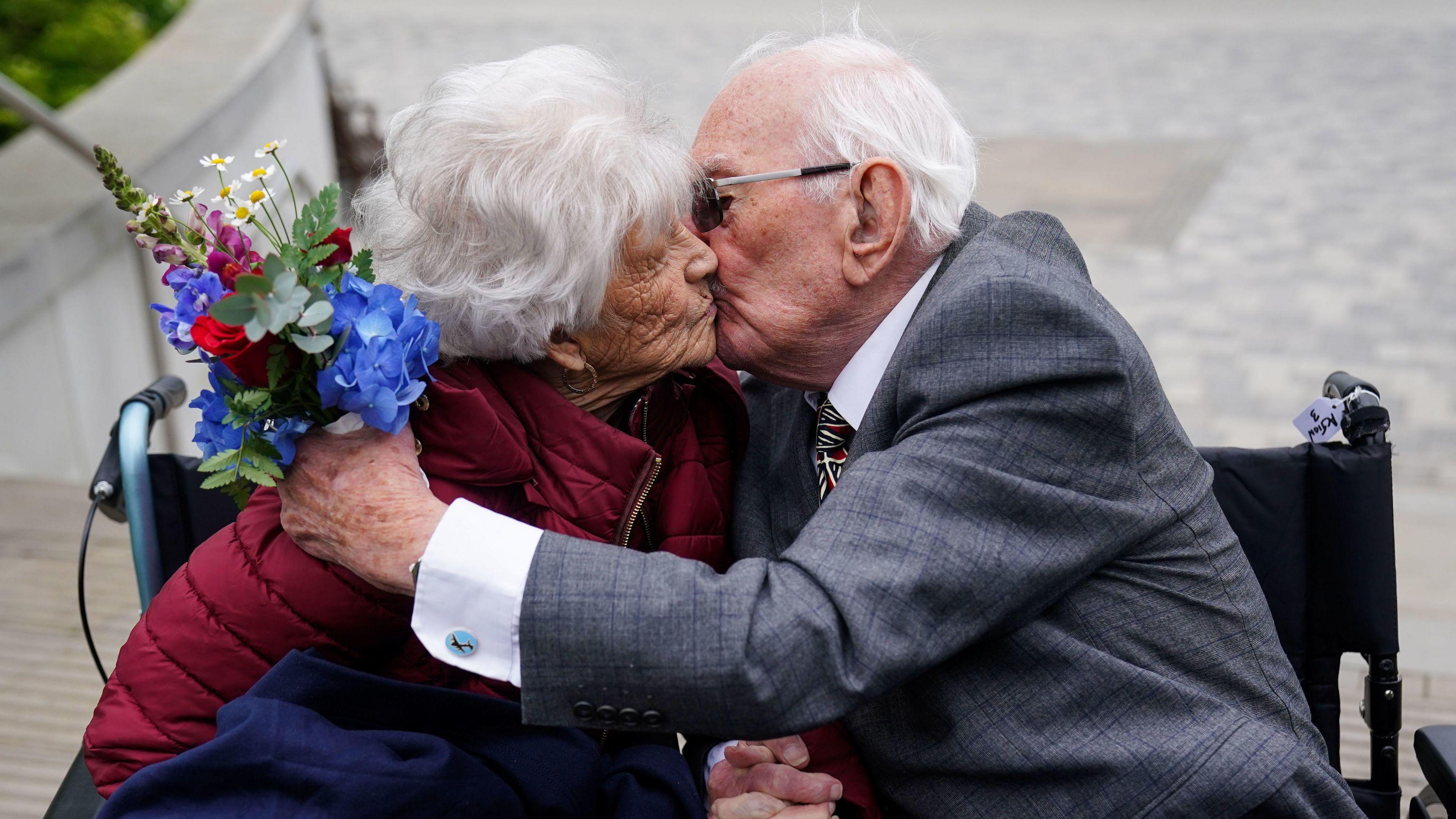 An elderly man and woman sitting in wheelchairs kiss as the man holds a bunch of flowers in his left hand.
