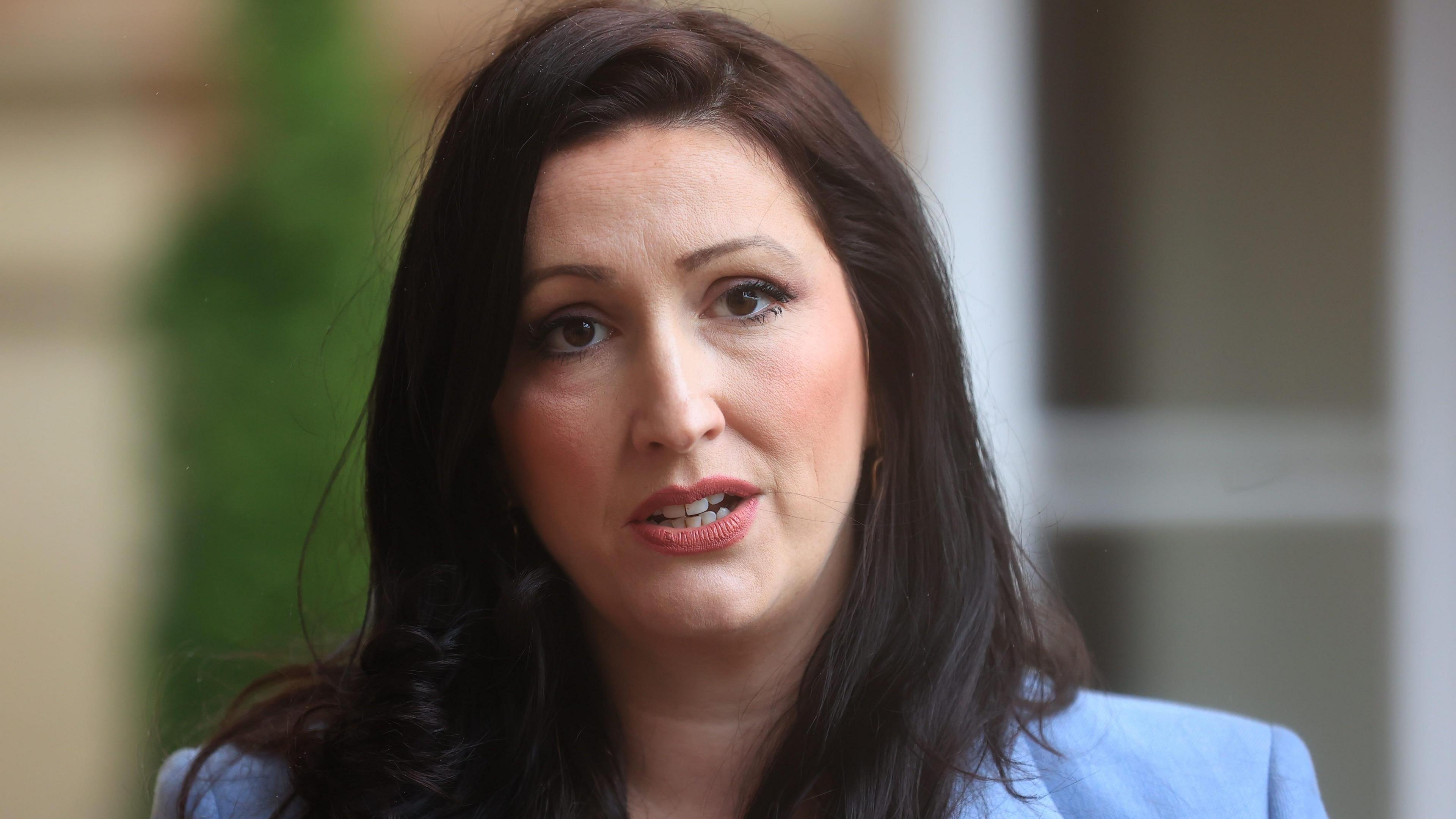 Emma Little-Pengelly wears a blue blazer. She has dark brown hair. The background behind her is blurred.