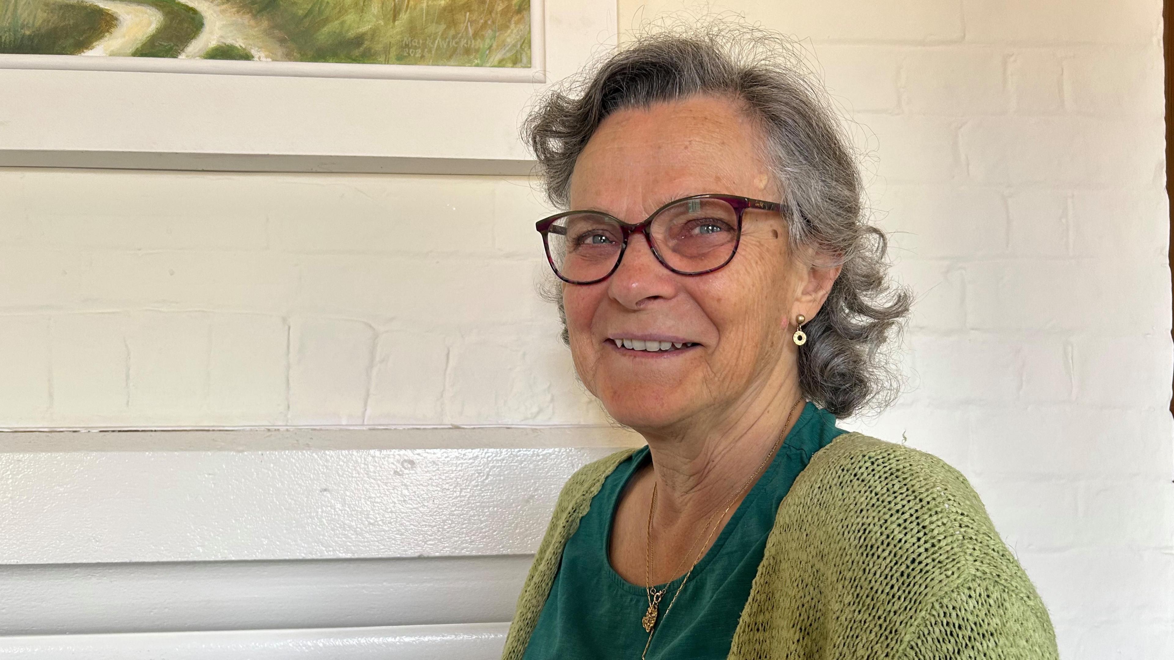 Kate Freeman smiles at the camera wearing a dark green top and light green cardigan. She wears a gold necklace and earrings. She has brown framed glasses on and short grey curly hair. The wall behind her is white painted brick.