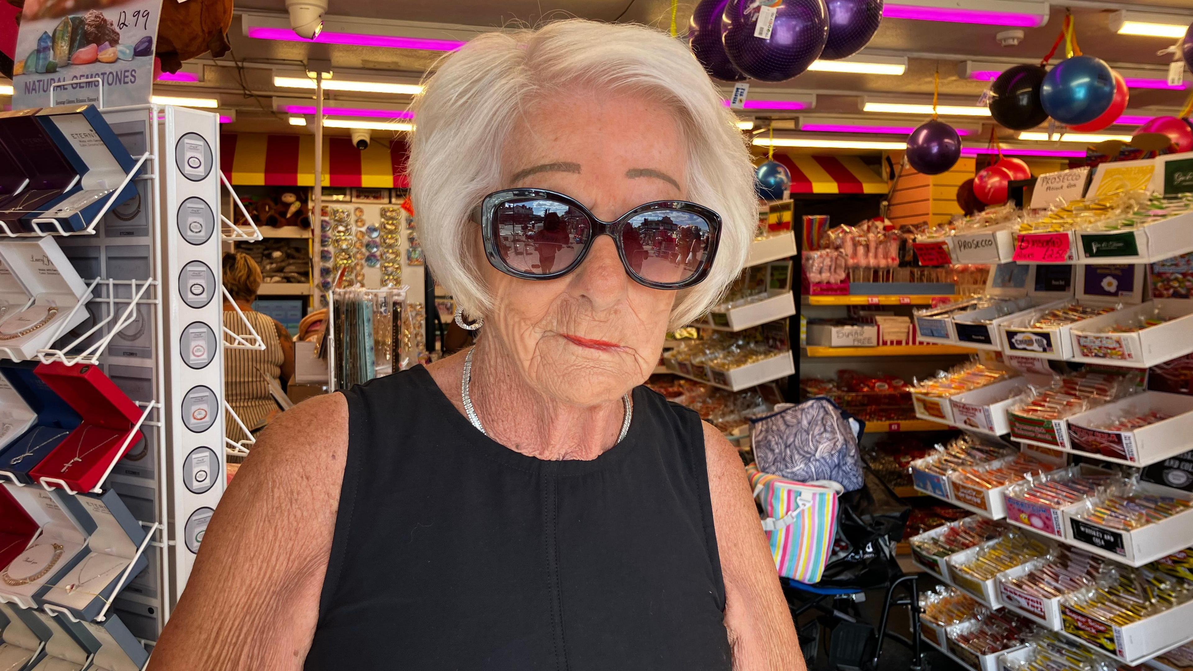 A woman in a black sleeveless dress looks at the camera. She has dark sunglasses and a silver necklace. She is standing in front of shelves full of sweets and toys.