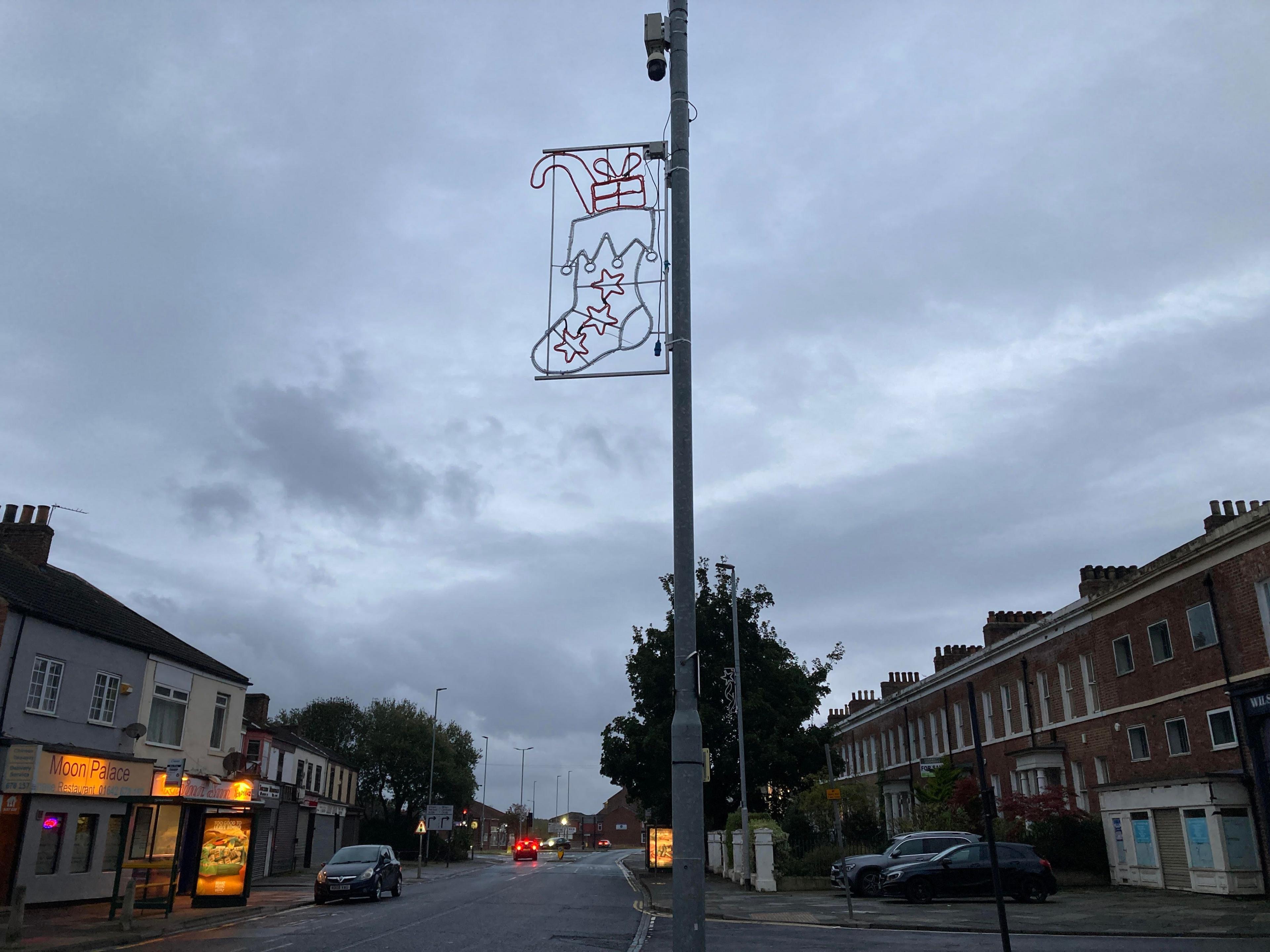 Christmas lights in the shape of a full Christmas stocking are attached to a CCTV post. A high street with various residences and shops can be seen in the background.