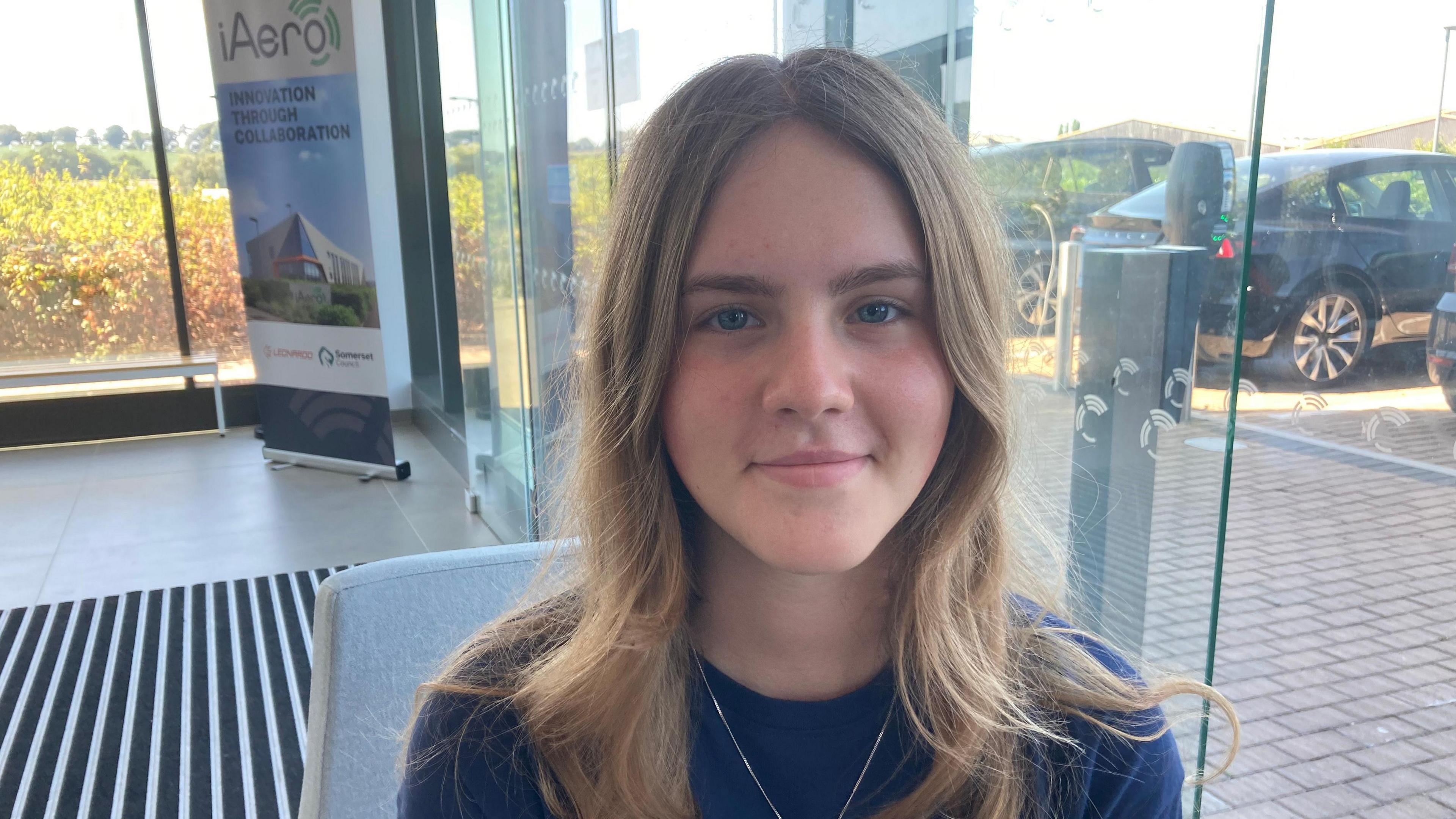 Girl with blonde hair looking at the camera. She's sat down on a chair by a glass panel window. There's a car park in the background. There's also a big A-board saying iAero on it.