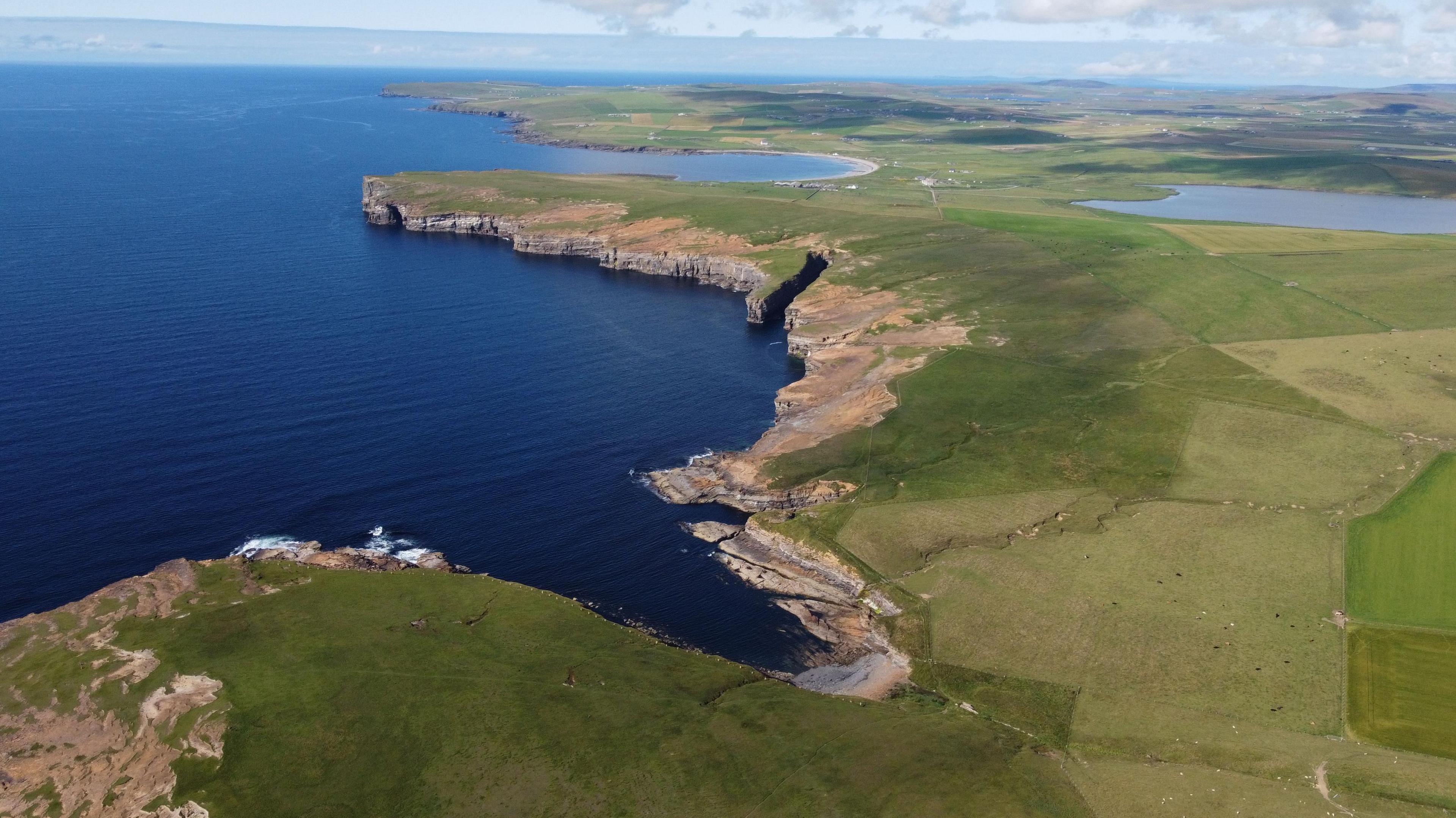 Aerial view of the rugged coastline of Orkney, with green fields and blue ocean under a partly cloudy sky.