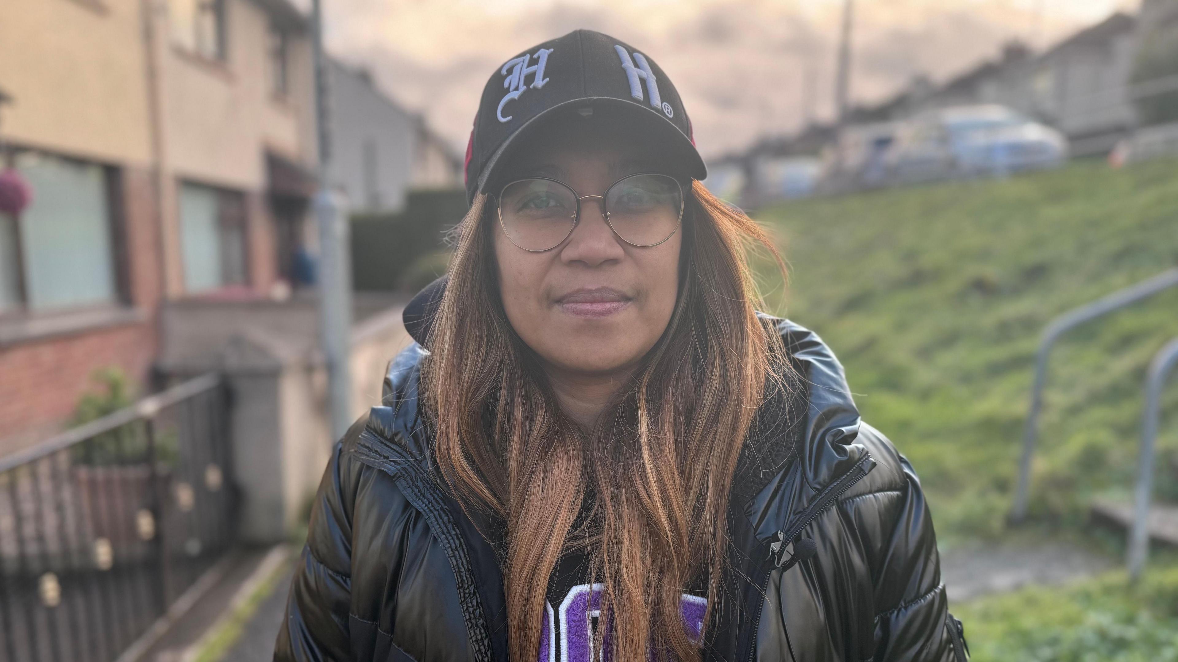 Cadalia Freitas - a woman with long, highlighted hair is wearing a baseball cap and circular glasses. She is also wearing a puffed black coat. She is standing on a footpath in front of a row of terraced houses.