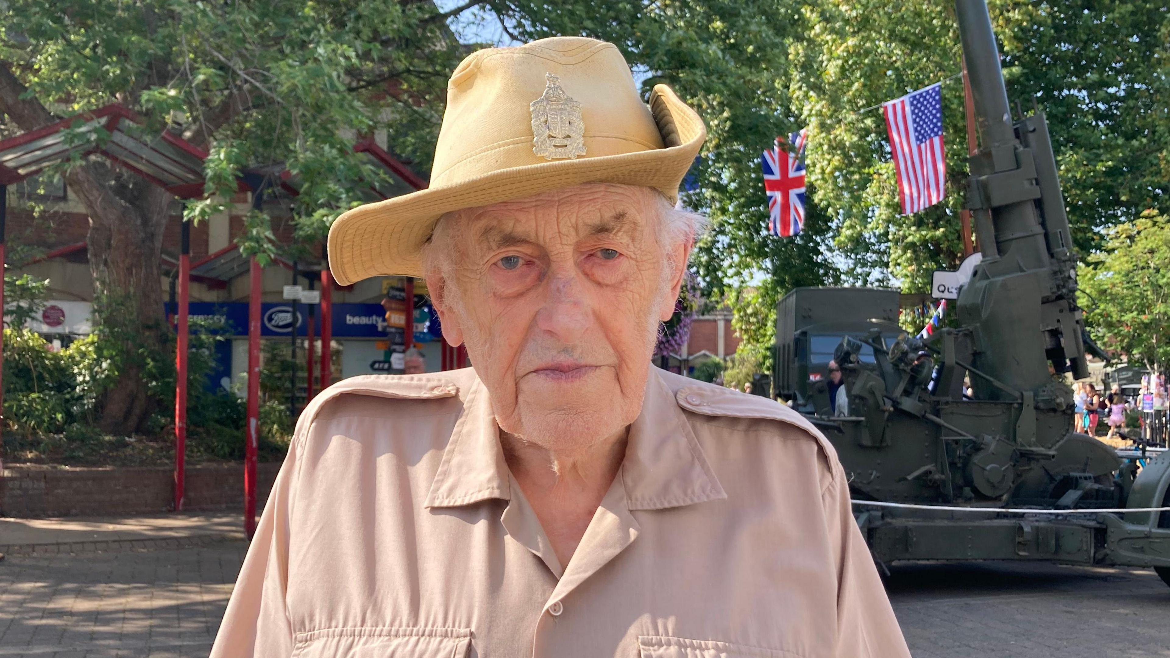 David Shopland looks at the camera with a serious expression. He wears a pale beige military shirt and a distinctive pale yellow hat with a wide rim and a military badge on the front. Behind him a camouflaged green military vehicle can be seen, as well as a Union Jack and the US flag hanging from a tree. 