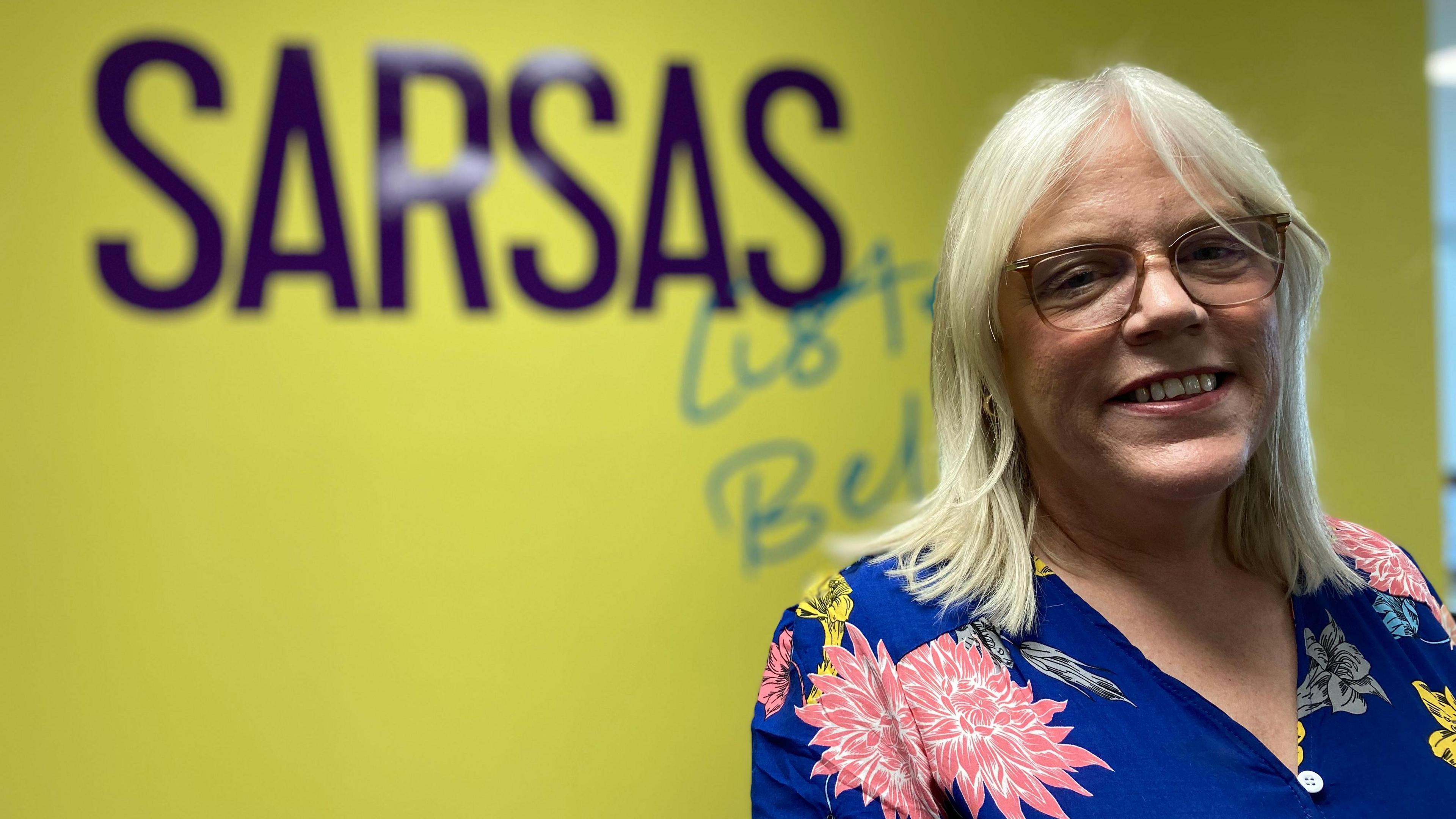 Lorri Weaving. She has shoulder length blonde hair and glasses. She is pictured in front of a yellow wall with the SARSAS logo printed on it. Lorri is smiling at the camera.