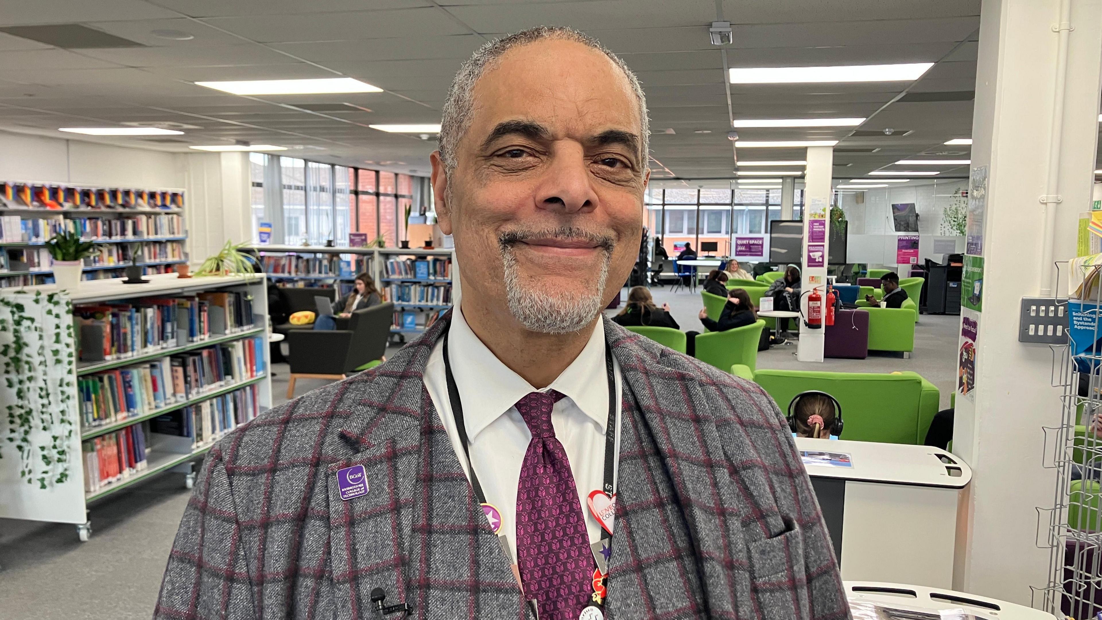 A man with grey hair and a grey beard wearing a tartan suit and smiling at the camera. He's standing in what appears to be a school common room with shelves of books behind him.