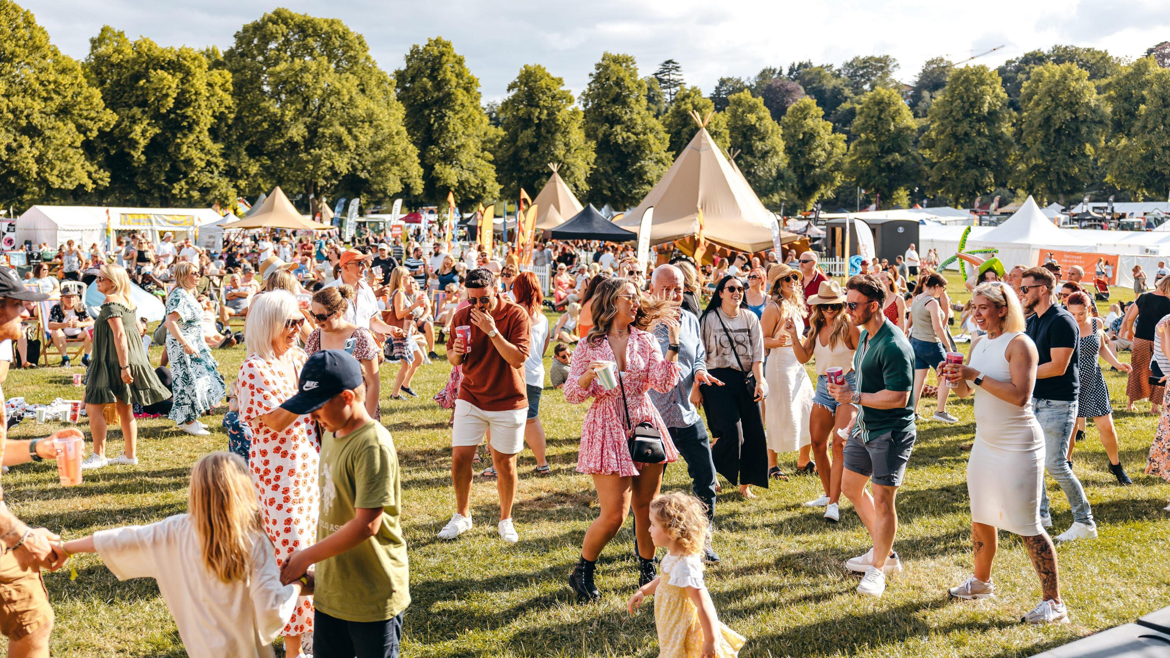 A large crowd of people on a green grass field. They are dancing and holding drinks. In the background are various tall tents and flagpoles. There is a line of trees in the background.
