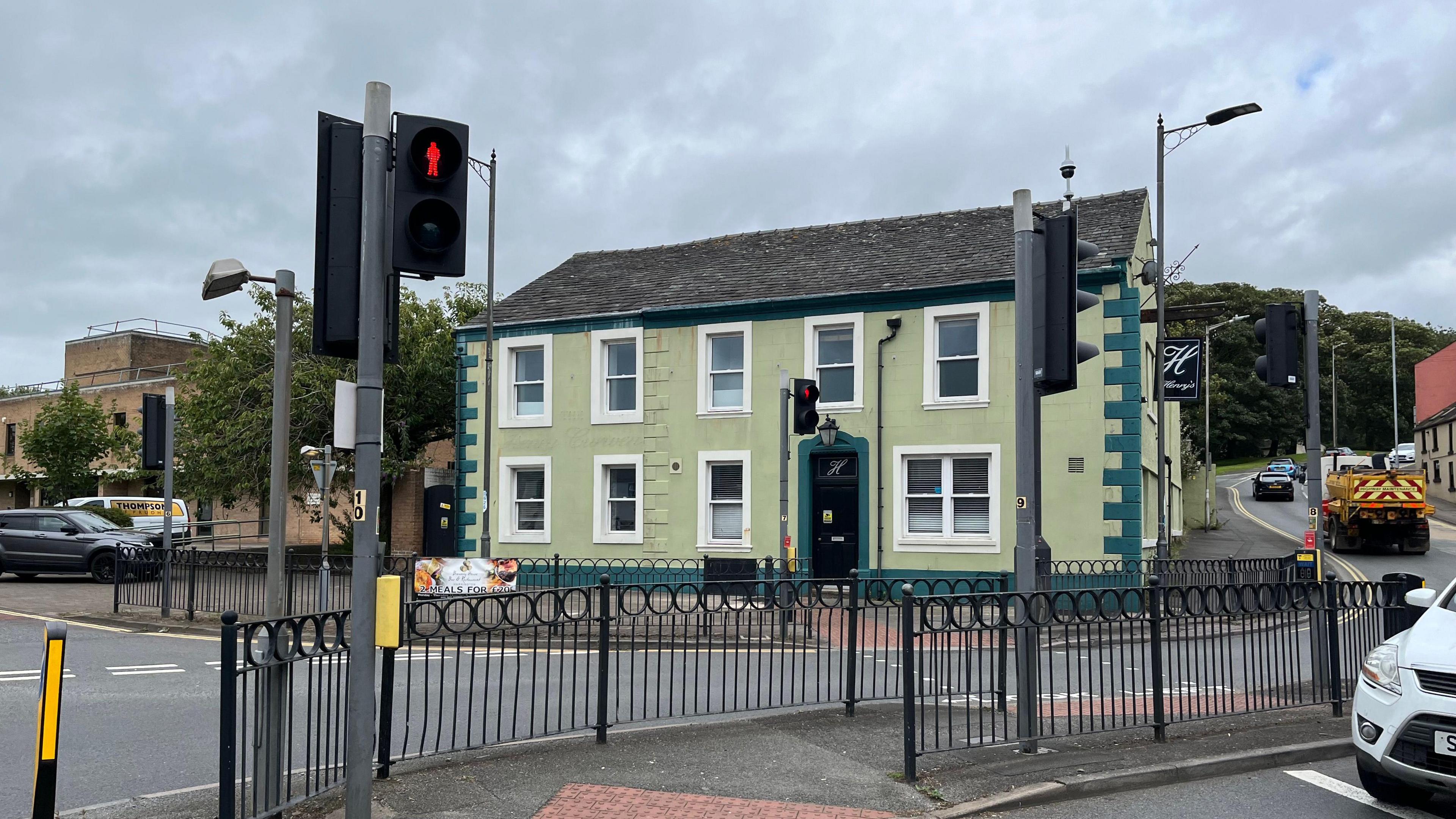 A big green house with nine windows on the side of a busy road. A pedestrian crossing can be seen in front of the building.