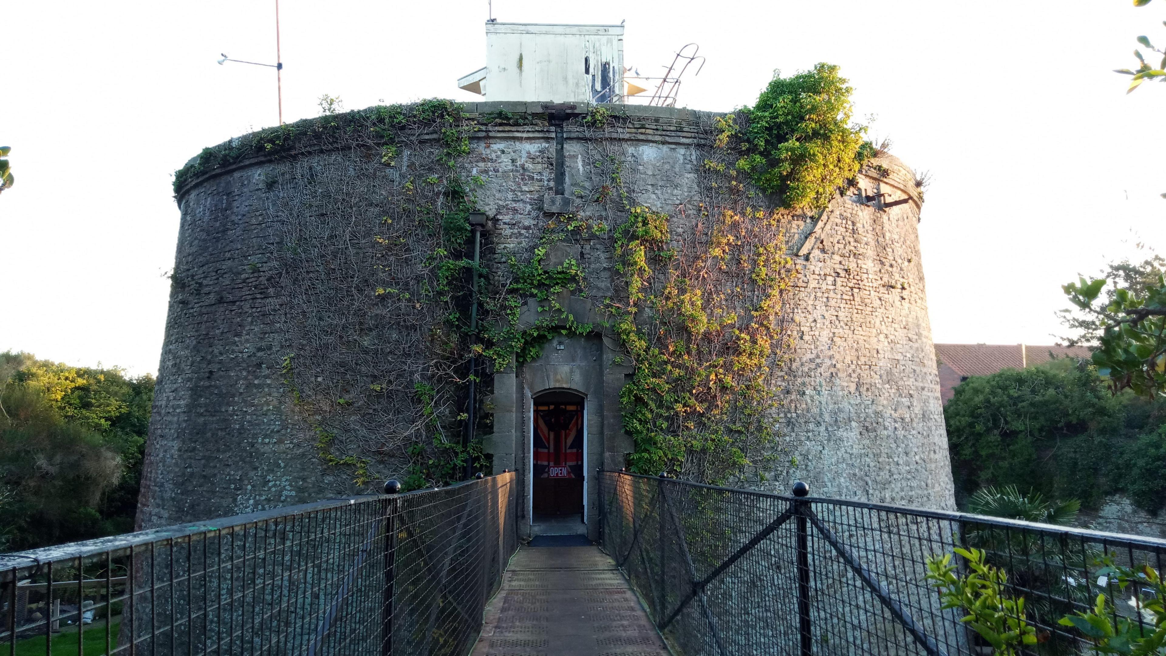 A cast-iron drawbridge leads towards the circular brick tower, which has ivy growing on part of its facade. A wooden door is at the end of the bridge.