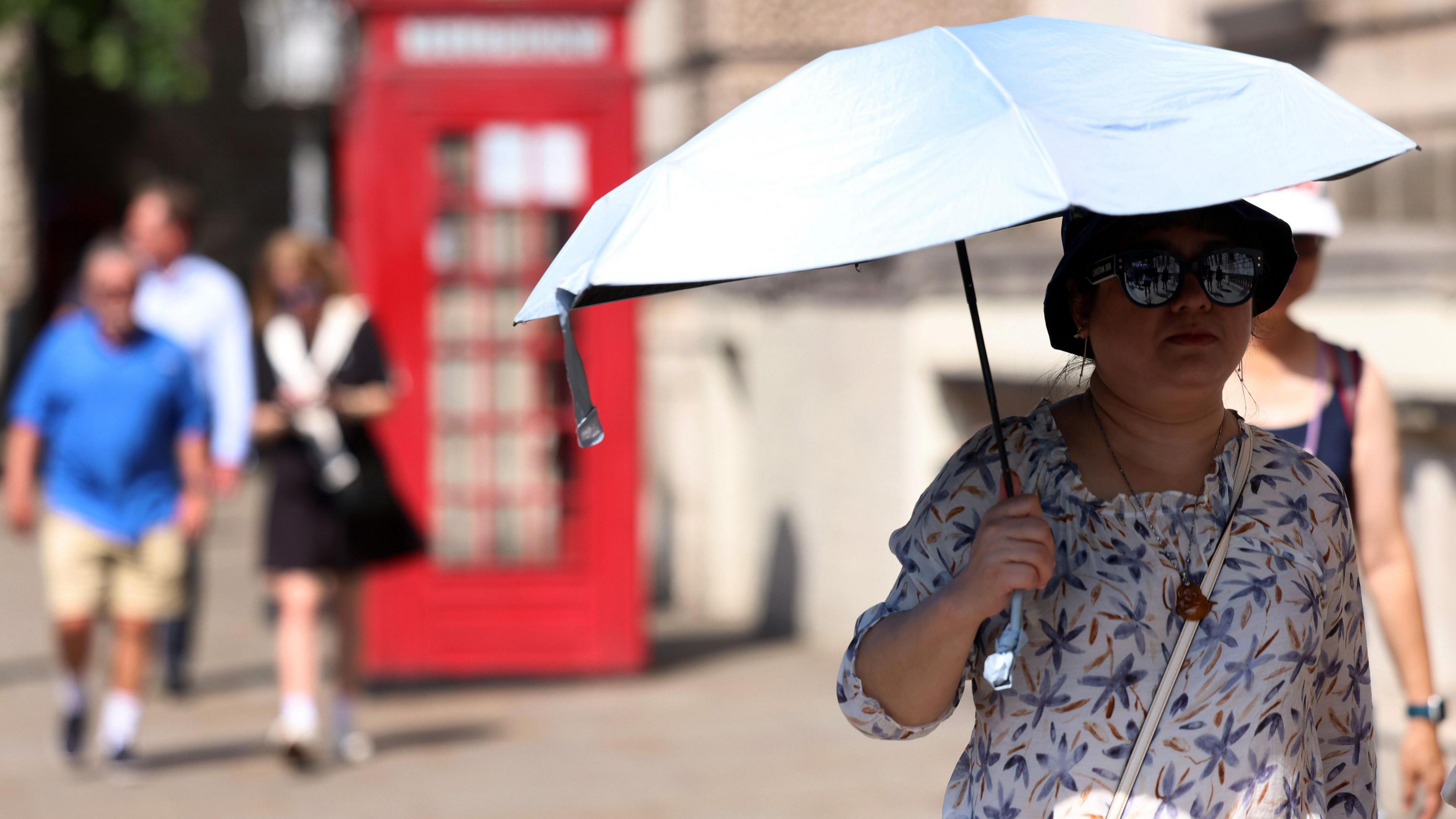A woman walks through a London street holding an umbrella to shield herself from the sun, with a red phone box and three other people in the background